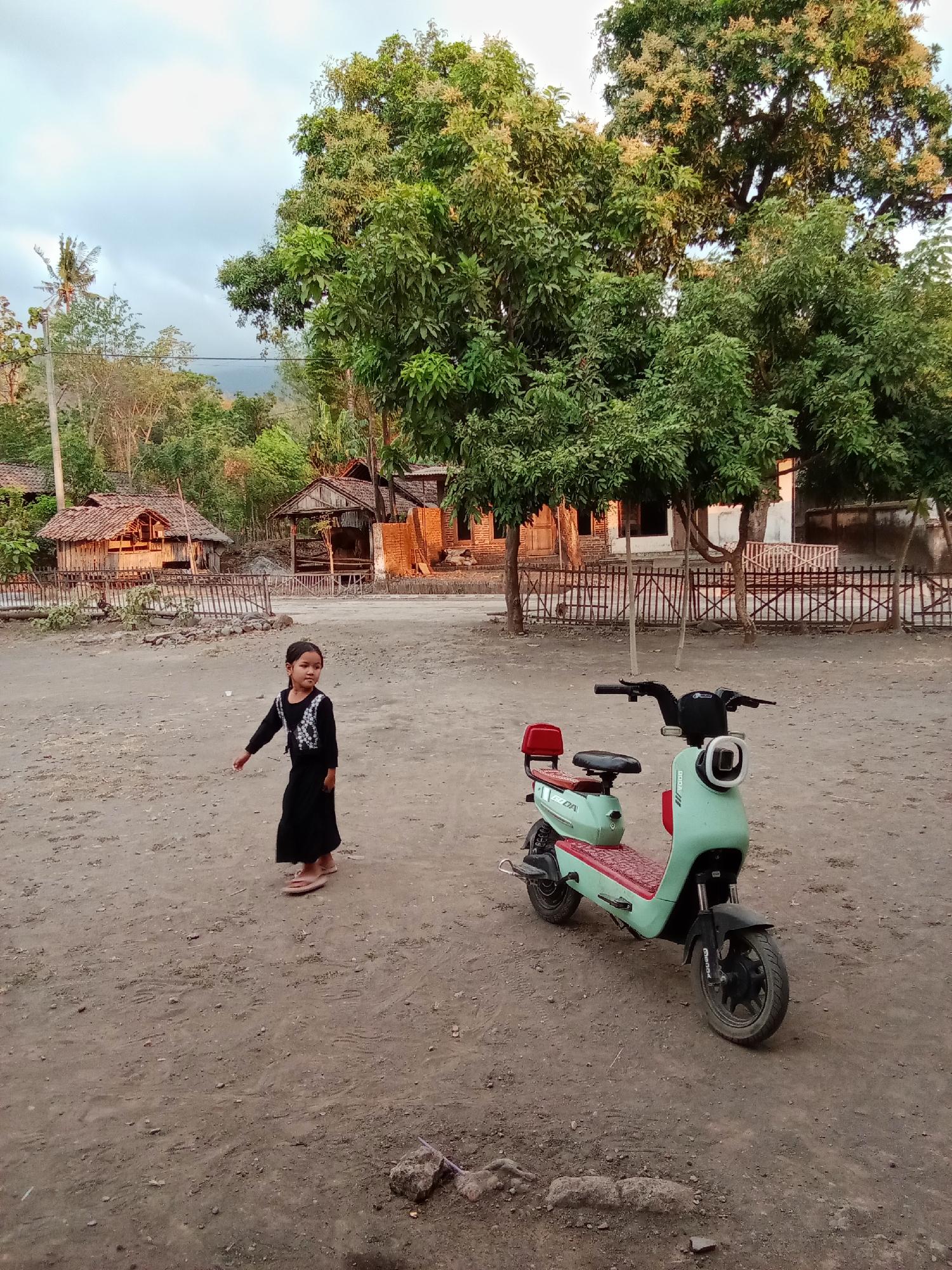 The image depicts a young girl in a black dress standing on a dirt ground in front of a light green and red electric scooter. Behind them, traditional houses with thatched roofs and wooden structures are visible, set against a backdrop of lush green trees and a cloudy sky. The scene appears to be an outdoor area in a rural village.
