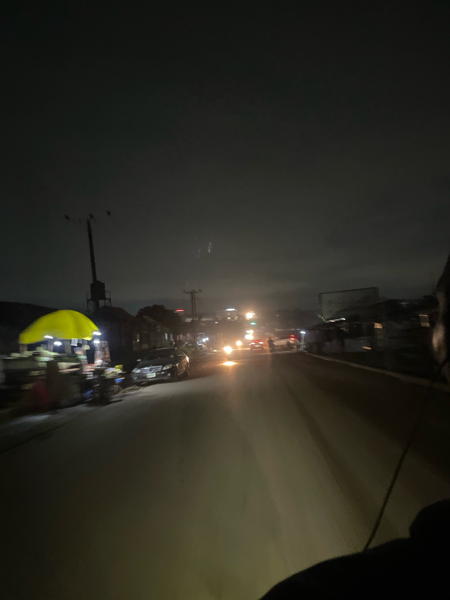 The image captures a nighttime scene on a street in Abuja, Nigeria, likely in a commercial or roadside vending area. The foreground is dominated by a dark road, partially illuminated by the headlights of passing vehicles. To the left, a roadside stall is visible, covered by a bright yellow umbrella. Underneath the umbrella, there are indistinct figures, possibly vendors or customers, and various items for sale. The stall appears to be lit, with small lights illuminating the products. Several cars are parked or driving on the road, their headlights creating streaks of light due to motion blur. In the background, there are buildings and structures, some with lights on, suggesting an urban or semi-urban environment. Utility poles with wires are also visible, characteristic of street infrastructure. The sky is dark, with some clouds that are faintly lit by the ambient city lights. The overall impression is that of a busy, active roadside at night in a Nigerian city, with commerce and traffic coexisting.