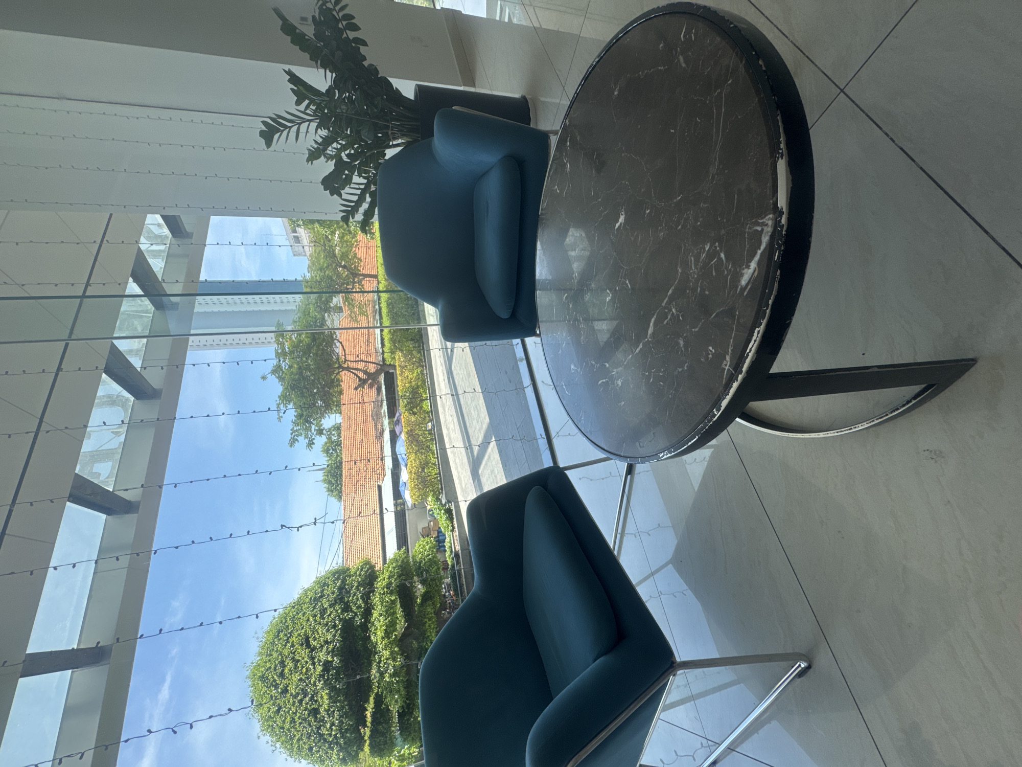 The image depicts a modern indoor seating area with two teal armchairs and a dark marble coffee table. These are situated in front of a large glass window that offers a view of lush greenery, red-tiled roofs, and a clear blue sky with wispy clouds. The interior features a tiled floor and what appears to be a tall potted plant to the right. String lights are visible hanging from the ceiling structure above the window. The scene is well-lit, suggesting it is daytime. The image does not contain any people or indicate specific actions or interactions.