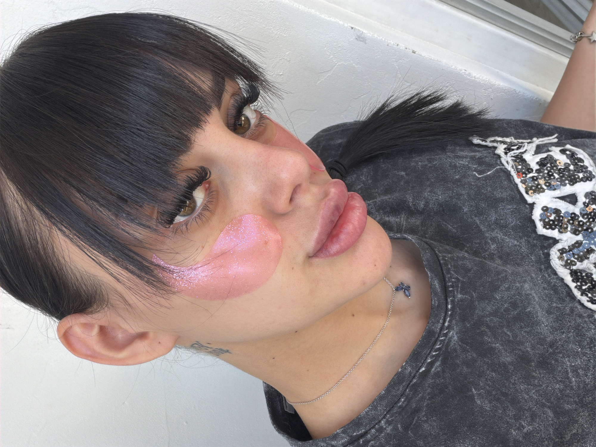 A young woman with dark hair, styled with bangs, and wearing a dark grey t-shirt with a butterfly necklace is pictured in a close-up, outdoor shot. She has prominent eyelashes and light pink, glittery under-eye masks. The background is a white wall. There is no indication of the city of Plovdiv, Bulgaria.