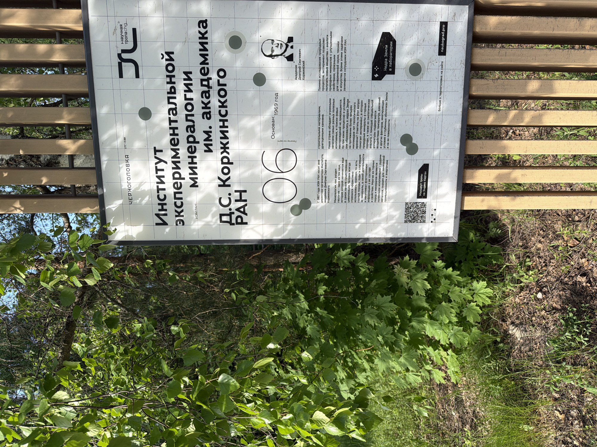 Here is a matter-of-fact description of the image:
The image shows a sign in the town of Chernogolovka, Russia, near Moscow. The sign provides information about the Institute of Experimental Mineralogy of the Russian Academy of Sciences (IEM RAN), named after Academician D.S. Korzhinsky. The sign notes that the institute was founded in 1969 and that, since 2017, IEM RAN has been named after its founding director, Academician Dmitry Sergeevich Korzhinsky. The sign explains the importance of mineralogy in geology and human history, and the role of the institute in studying the formation of minerals and developing new materials. There is a QR code to provide more information.
