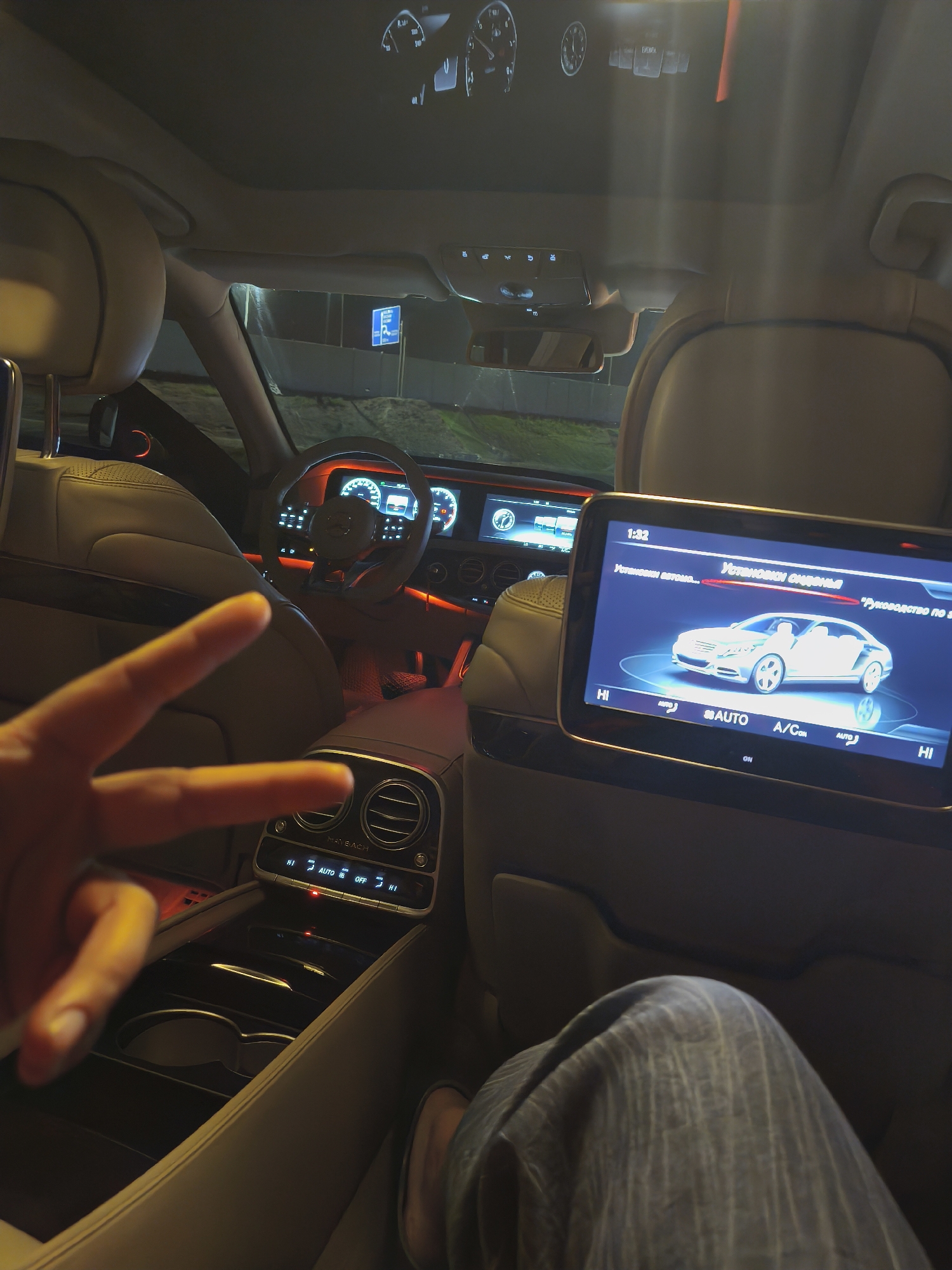 The image shows the interior of a luxury car, likely a Maybach, at night, possibly in the vicinity of Сокуры, Russia, given the Cyrillic script on the screen. The view is from the backseat, with a screen displaying car settings, including the climate control and seat adjustment options. The car's interior features light-colored leather upholstery and ambient lighting, with a dashboard and steering wheel visible in the background. A hand is partially visible, making a peace sign. Part of a person's leg is also in the foreground. The car appears to be parked, as indicated by the visible street sign.