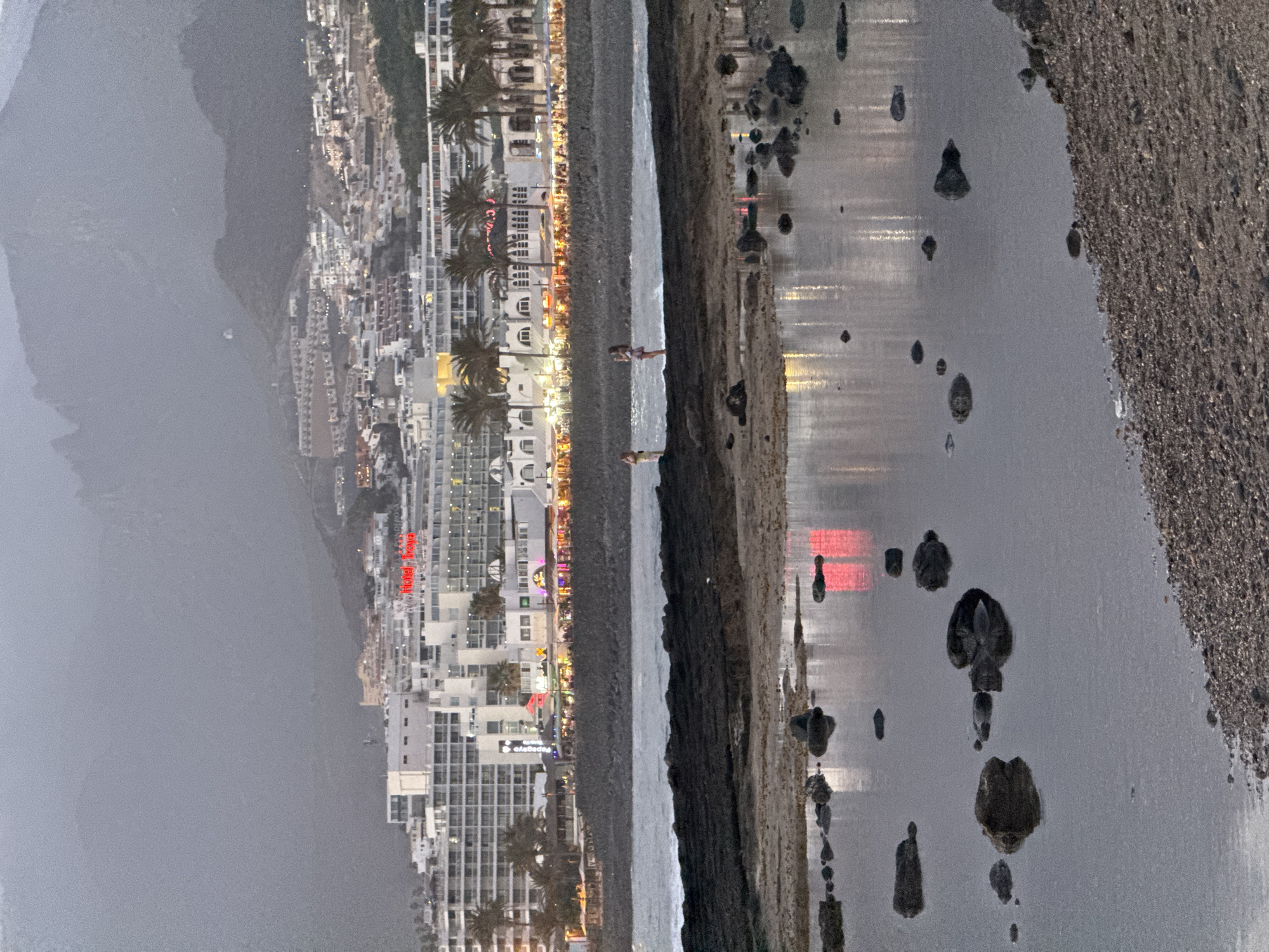 The image depicts a coastal scene at dusk in Arona, Spain. In the foreground, a shallow body of water, likely tide pools, reflects the lights from the town. Rocks of various sizes are scattered within and around these pools. The middle ground shows a rocky shoreline with two individuals standing on the rocks, facing the water. Beyond the shoreline, a promenade is lined with buildings, palm trees, and brightly lit establishments, indicating a commercial and tourist area. The background features a large, multi-story hotel complex and further in the distance, a mountain range shrouded in mist or clouds. The overall lighting suggests it is either early morning or late evening, with artificial lights from the town illuminating the scene. The architecture of the buildings suggests a Mediterranean or resort style. The sea is calm with small waves lapping at the shore.