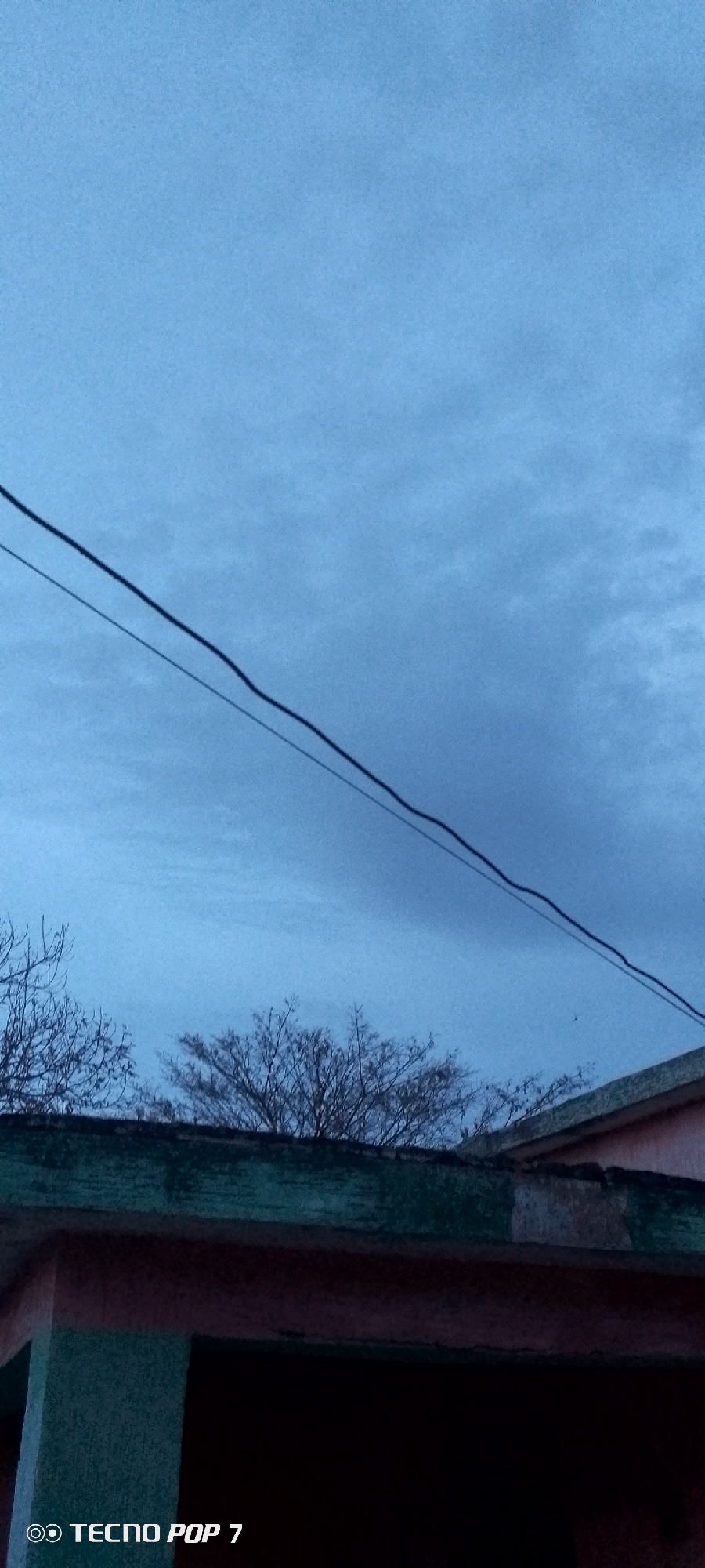 The image depicts an overcast sky in Altagracia, Venezuela. In the foreground, there is a building with a roof and some trees. Overhead, two parallel electrical wires stretch across the frame. The "TECNO POP 7" watermark is visible in the lower left corner.