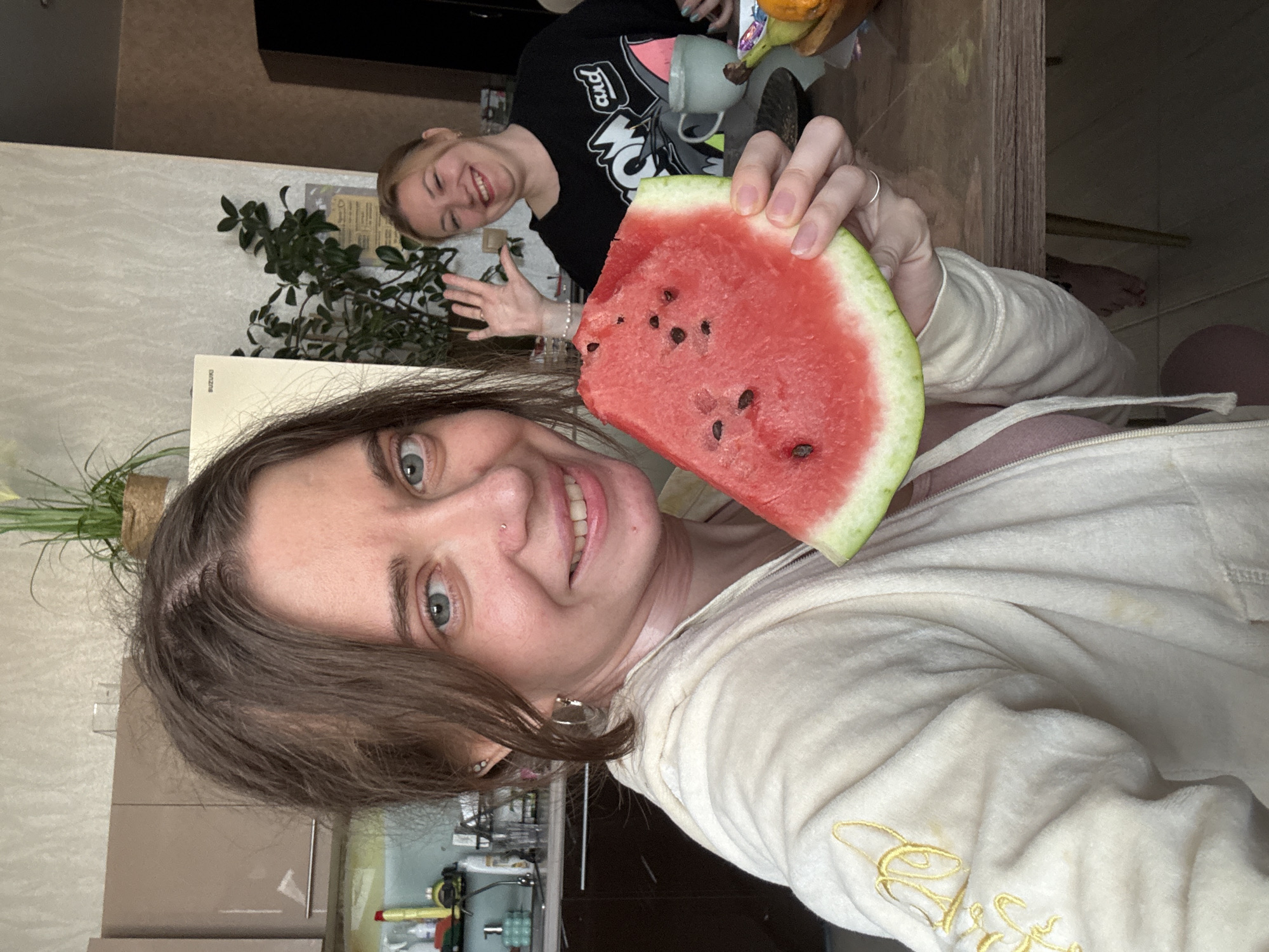 Two women are present in a kitchen. The woman in the foreground is holding a slice of watermelon and looking at the camera with a smile. She is wearing a light-colored hooded jacket and a pink top. She has light brown hair and blue eyes and is wearing hoop earrings and a nose ring. The woman in the background is smiling and waving, wearing a black sweatshirt. She has blonde hair. The kitchen has wooden cabinets, a countertop with various items, and a table where the women are seated. A plant is visible in a pot on a shelf in the background.