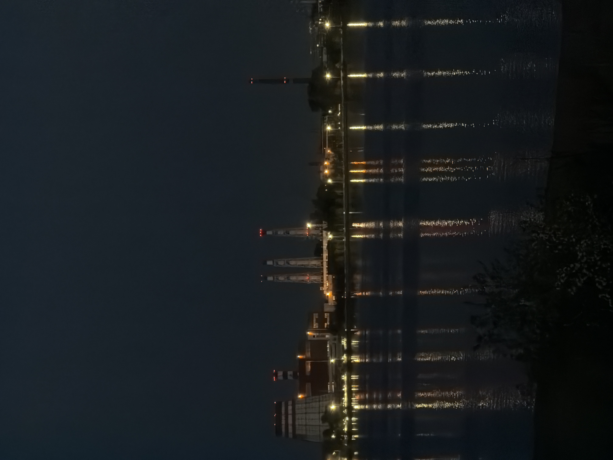 The image shows a nighttime view of a river or lake in Izhevsk, Russia, reflecting lights from buildings and infrastructure along the shoreline. Visible structures include industrial buildings with chimneys, cranes, and streetlights. The city lights are reflected in the water, creating streaks of light across the surface. Some vegetation, possibly trees or bushes, is visible along the immediate shoreline.