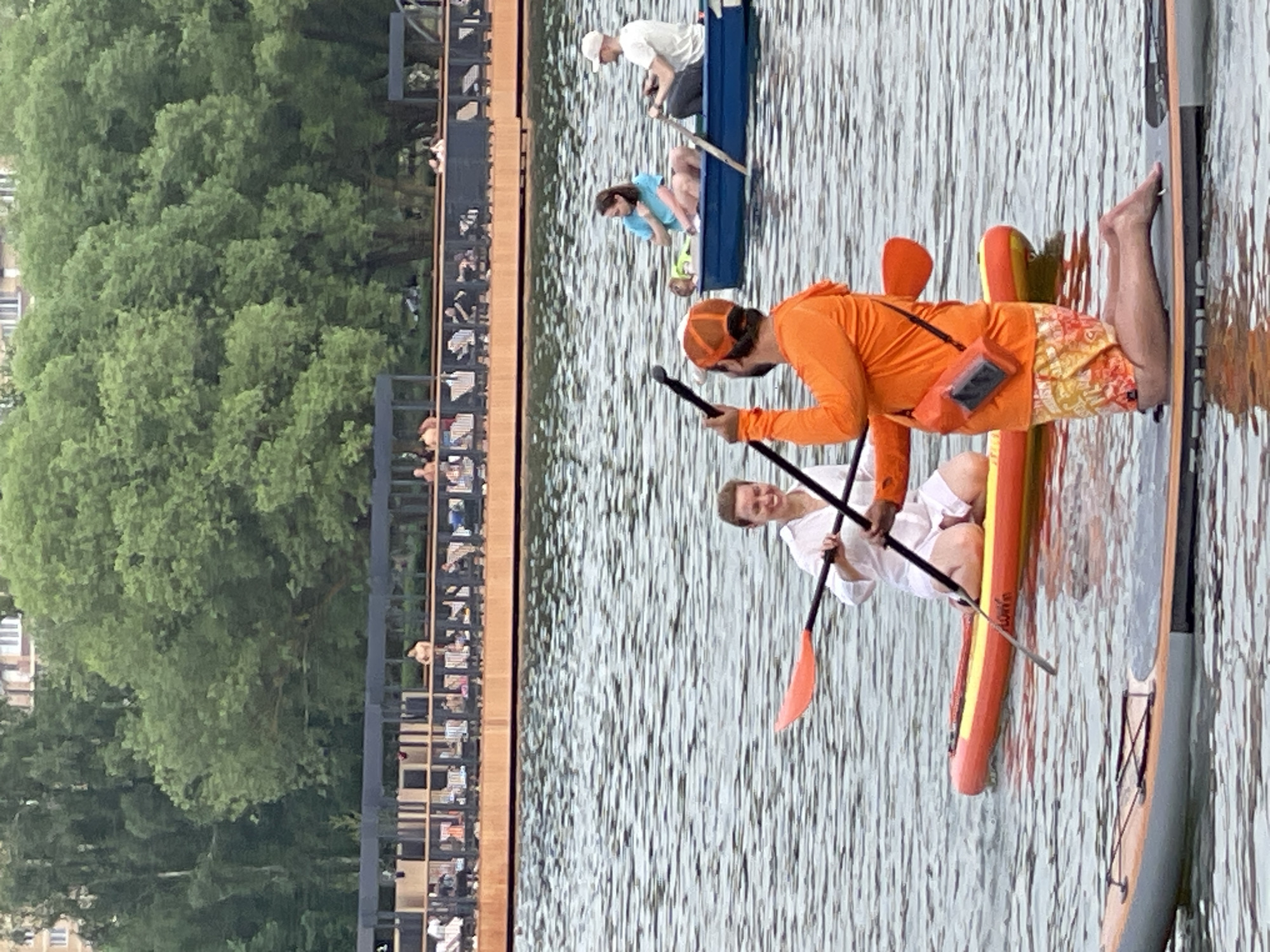 In Moscow, Russia, on a body of water, two individuals are engaged in paddleboarding. In the foreground, a person wearing an orange hooded sweatshirt and patterned shorts kneels on a paddleboard, holding a paddle. To their left and slightly behind, a woman in a white shirt sits on another paddleboard, also holding a paddle. In the background, a blue boat is on the water, with three people inside: two rowing and one woman in a blue shirt. Further back, a wooden structure with railings and people sitting on it runs along the waterfront, backed by a dense line of green trees. The water is calm with ripples.