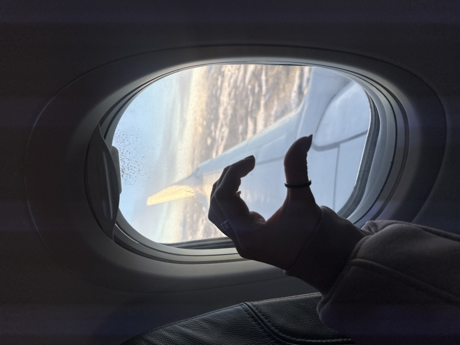 The image depicts an interior view from an aircraft, with an implied location above Teigebyen, Norway. A human right hand, adorned with a ring on the ring finger and a dark band on the pinky, is positioned in the foreground, forming a 'half-heart' gesture. The hand is partially covered by a light-colored, long-sleeved garment with a ribbed cuff. This hand is placed against the oval-shaped airplane window frame. Through the window, a white aircraft wing is visible, extending diagonally from the mid-left towards the lower right. The external environment features a sky gradient from light blue at the top to yellow and orange hues near the horizon, indicative of either sunrise or sunset. Below this, a dense, textured layer of clouds or snow-covered terrain is visible. The upper right portion of the windowpane exhibits small, crystalline formations, likely ice or condensation. A dark, upholstered airplane seatback is partially visible on the left side of the frame.