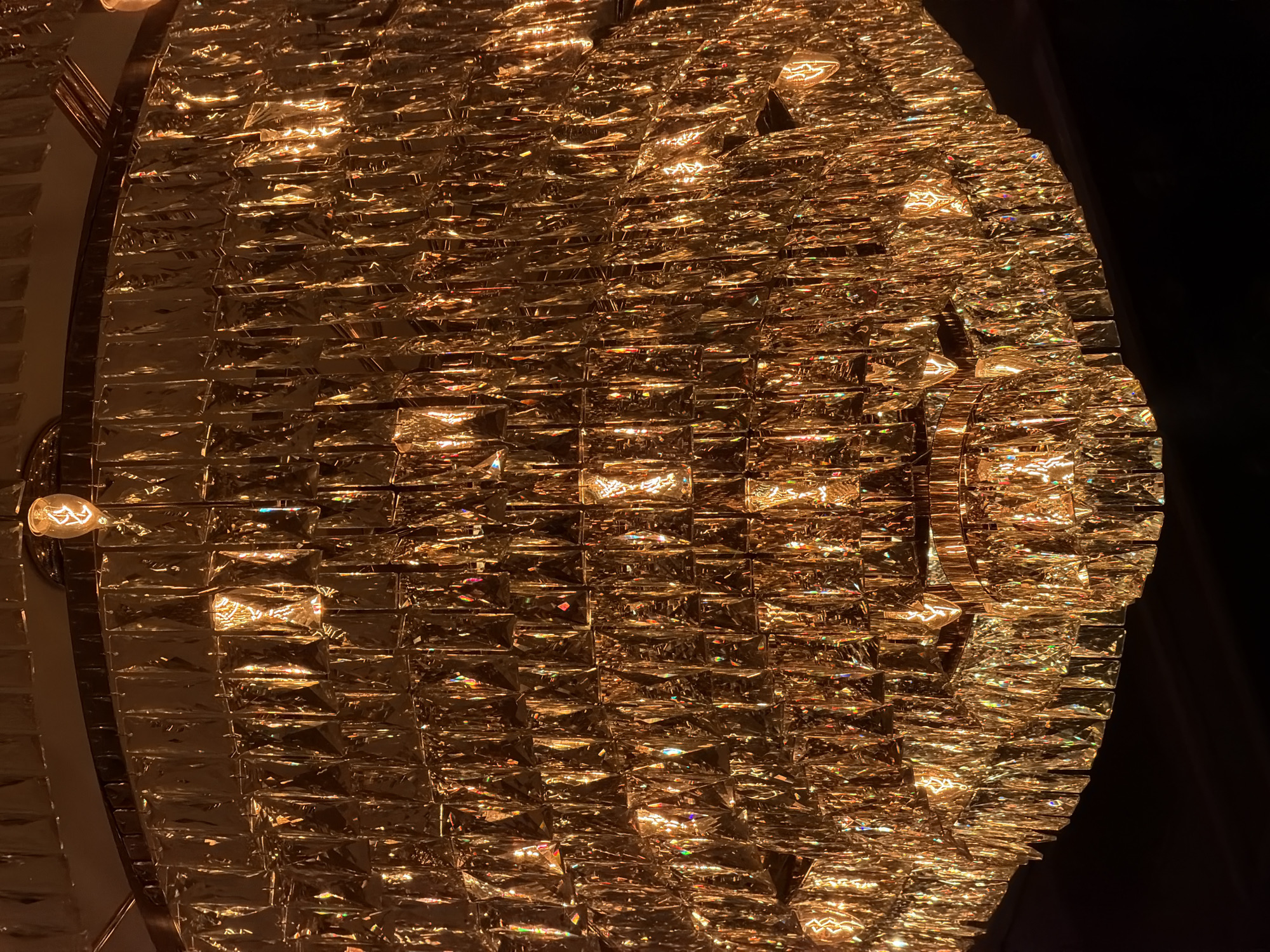 The image shows a close-up view of a decorative lighting fixture, possibly a chandelier, composed of numerous small, rectangular, reflective elements. These elements are arranged in a grid-like pattern, creating a shimmering effect with various light sources visible within the fixture. The color palette is dominated by warm tones of gold and amber. The overall shape of the fixture appears to be circular or oval. There are also exposed light bulbs visible around the edge of the fixture. The background is dark, providing contrast and emphasizing the illuminated nature of the subject. There is no way to know if it is in Moscow, Russia from the image alone.