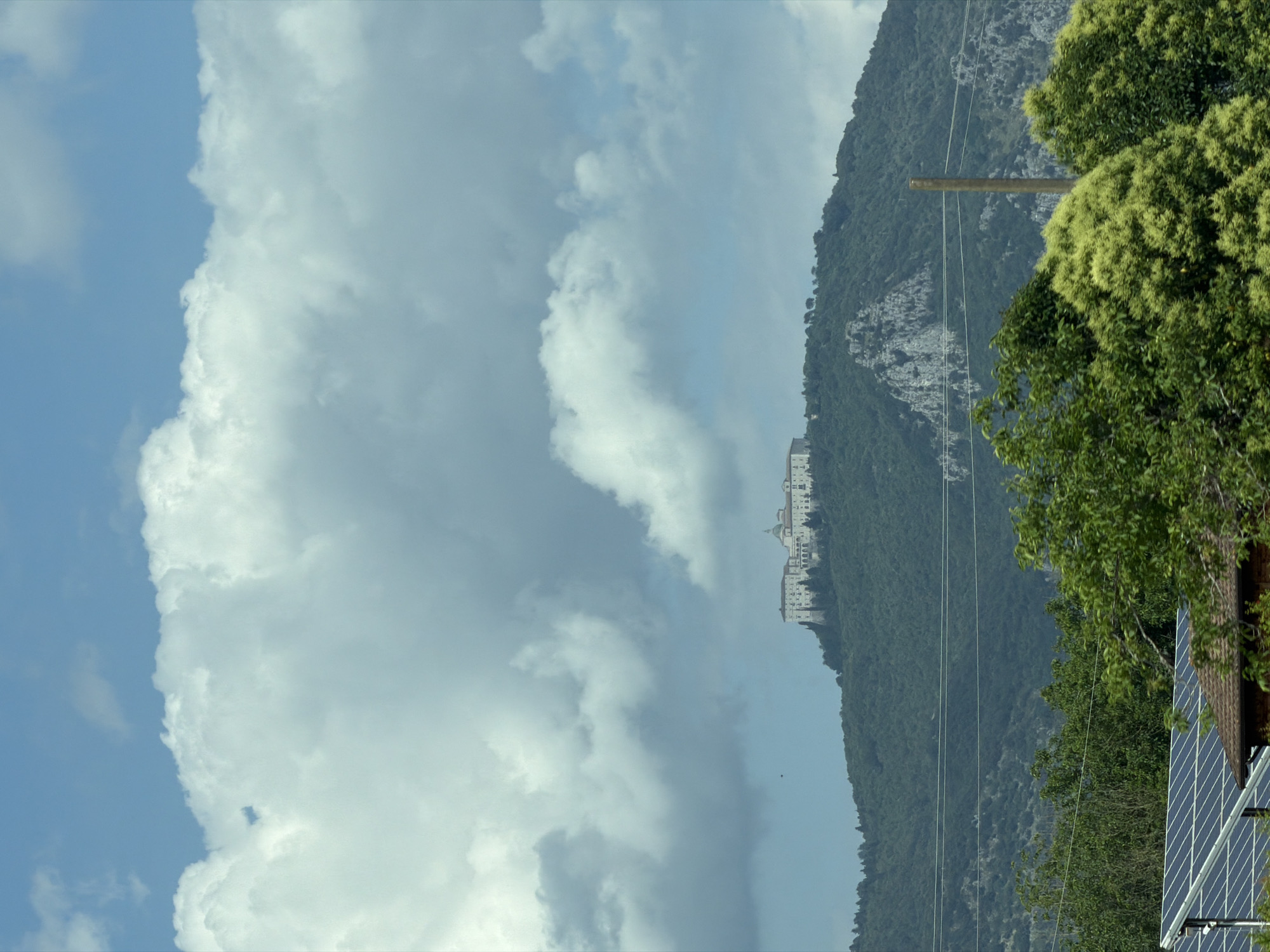 The image, correctly oriented, captures a panoramic landscape featuring mountains, sky, and a prominent building. The upper portion of the frame is dominated by a bright blue sky filled with large, white, and fluffy cumulus clouds, creating a dynamic sky scene.

In the mid-ground, a verdant mountain range stretches across the horizon, covered extensively in lush green trees and foliage. Rocky outcrops are visible along the mountain's contours. Perched majestically atop a distant, higher peak on the mountain is a large, ornate white building complex, featuring multiple stories, domes, and numerous windows. This structure strongly resembles the historic Abbey of Monte Cassino, fitting the provided location context of Piedimonte San Germano, Italy.

In the foreground, on the right side, dense green trees with lighter green, possibly flowering, foliage partially frame the scene. Below these trees, in the bottom right corner, a section of a reddish-brown tiled roof equipped with blue solar panels is visible. A wooden utility pole and several thin power lines extend horizontally across the mid-ground on the right side, running in front of the mountain.

The scene depicts a bright, clear day, likely late morning or early afternoon, with natural light illuminating the landscape. There are no people visible, and no specific activities are taking place. No text is discernible in the image.