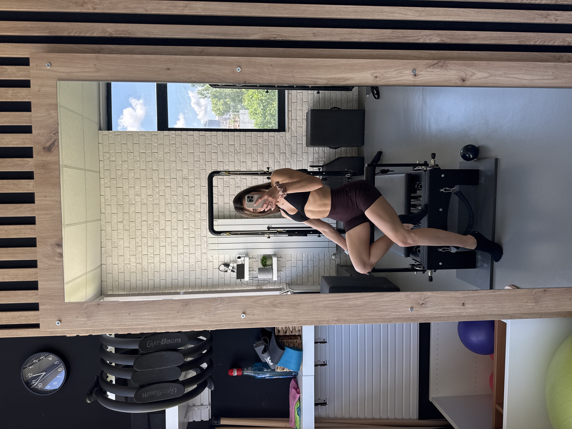 A young woman with brown hair, wearing a black sports bra and dark brown shorts, is taking a mirror selfie in a well-lit gym. She is sitting on a Pilates reformer machine. The gym has white brick walls, and a large window in the background reveals a cityscape with green trees and blue sky with clouds. To the left of the reformer, there is a shelf with a blue bottle, a basket with resistance bands, and cleaning cloths. Above the shelf, a circular black clock with a white face is mounted on a black wall. Several black Pilates rings are hanging on a rack to the left of the clock. To the right of the mirror, vertical wooden slats are installed on the wall. In the lower left corner, storage units are visible, containing a purple exercise ball and other gym equipment. The floor is gray. The overall impression is that of a modern, clean, and well-equipped fitness studio. This image may have been taken in Sofia, Bulgaria.