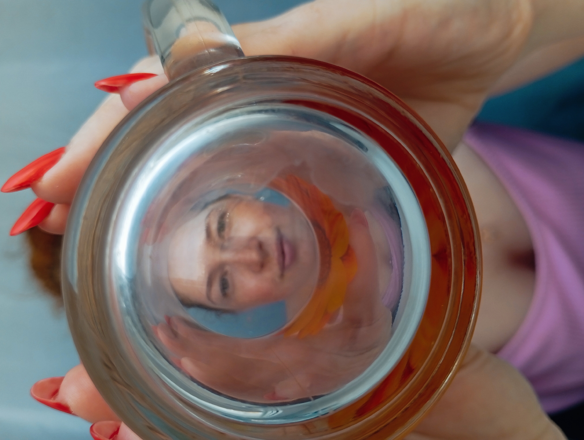 A woman's face is seen through the bottom of a clear glass mug held by two hands with long red fingernails. The woman's reflection is distorted by the glass, showing a slightly blurred view of her face, an orange scarf or piece of clothing, and a pink shirt. The background is indistinct, with hints of blue. The location cannot be determined to be Yurgamysh, Russia.