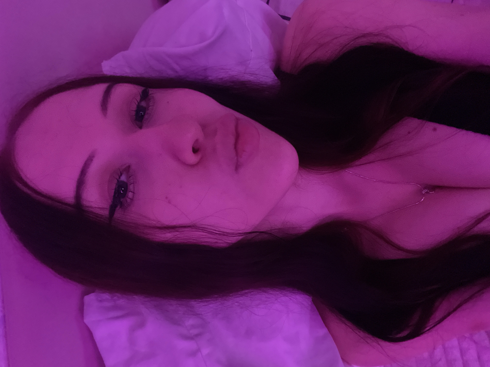 The image shows a close-up, selfie-style portrait of a young woman with long, dark hair, wearing a necklace. She is lying down on a pillow, with a pink light illuminating the scene. Her eyes are looking towards the camera, and her lips are slightly pursed. The background appears to be a bed or sofa. There is no discernible information about the location being Votkinsk, Russia, or any specific environmental details that would indicate such a location. The image focuses solely on the subject.
