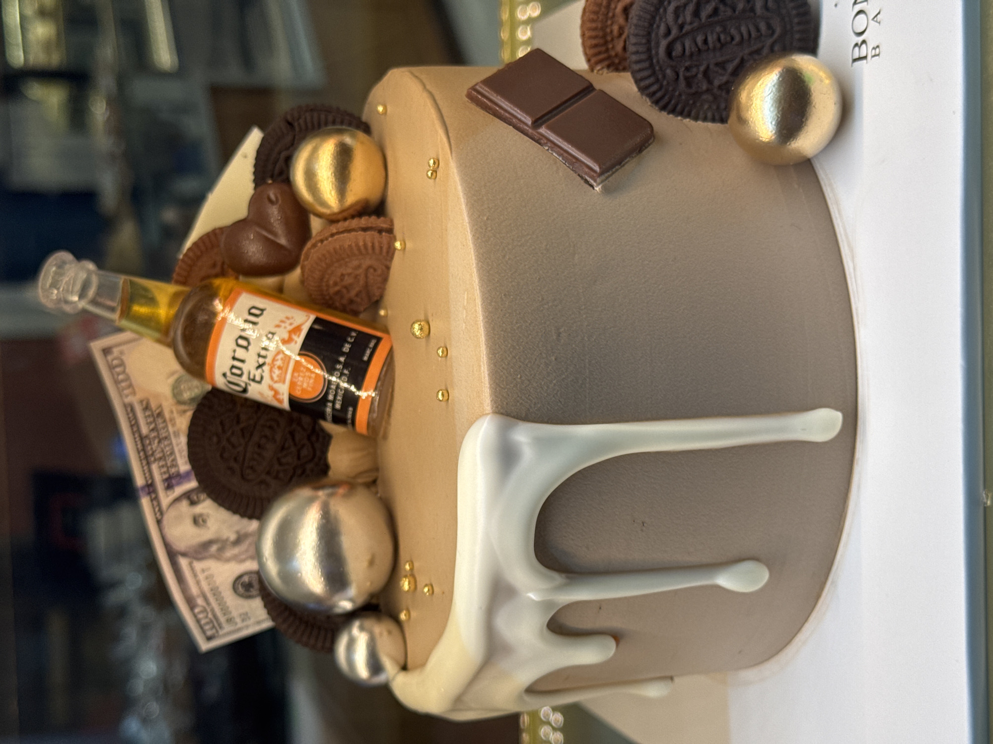 A chocolate cake with white drip frosting and a miniature Corona Extra beer bottle, chocolate cookies, gold and silver spheres, a chocolate bar, and fake money adorns a white cake board. The cake is on display in a bakery. The background is blurred, showing shelves stocked with baked goods. The logo on the cake board appears to be "BON BAKE".