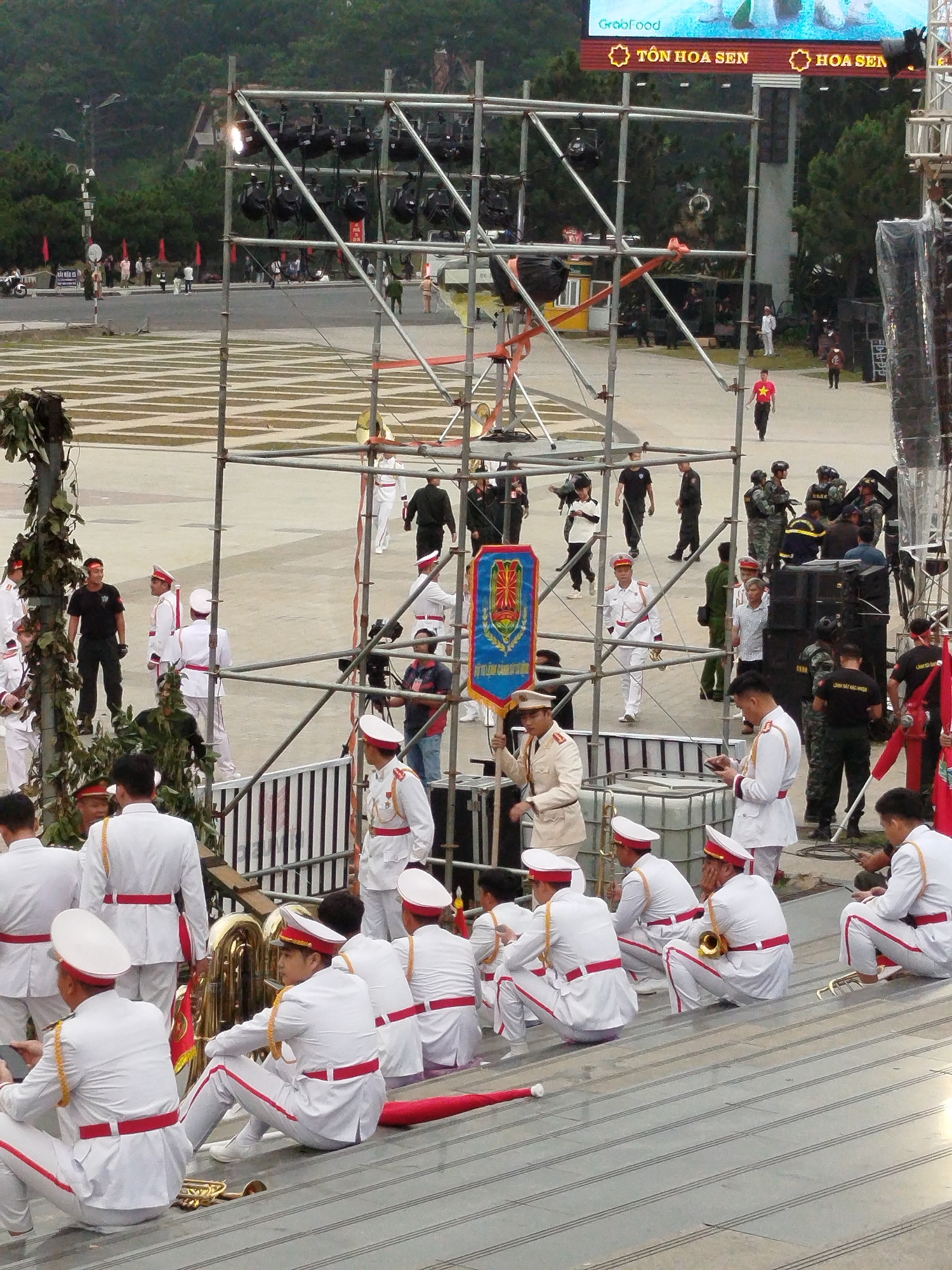 The image depicts an outdoor event in Da Lat, Vietnam, likely a ceremony or performance. The foreground features several individuals in white military-style uniforms with red sashes and trim, some seated on tiered steps, others standing. Some are holding musical instruments, including a tuba and a brass instrument, suggesting a band is present. Behind them, a large metal scaffolding structure is erected, supporting multiple spotlights. Various other individuals in different attire, including some in black and camouflage uniforms, are visible in the midground and background, mingling and observing. The setting includes a paved area with marked grids, a large screen displaying text and images, trees, and a hilly landscape in the distance. The event appears to be taking place in an open plaza or public square.