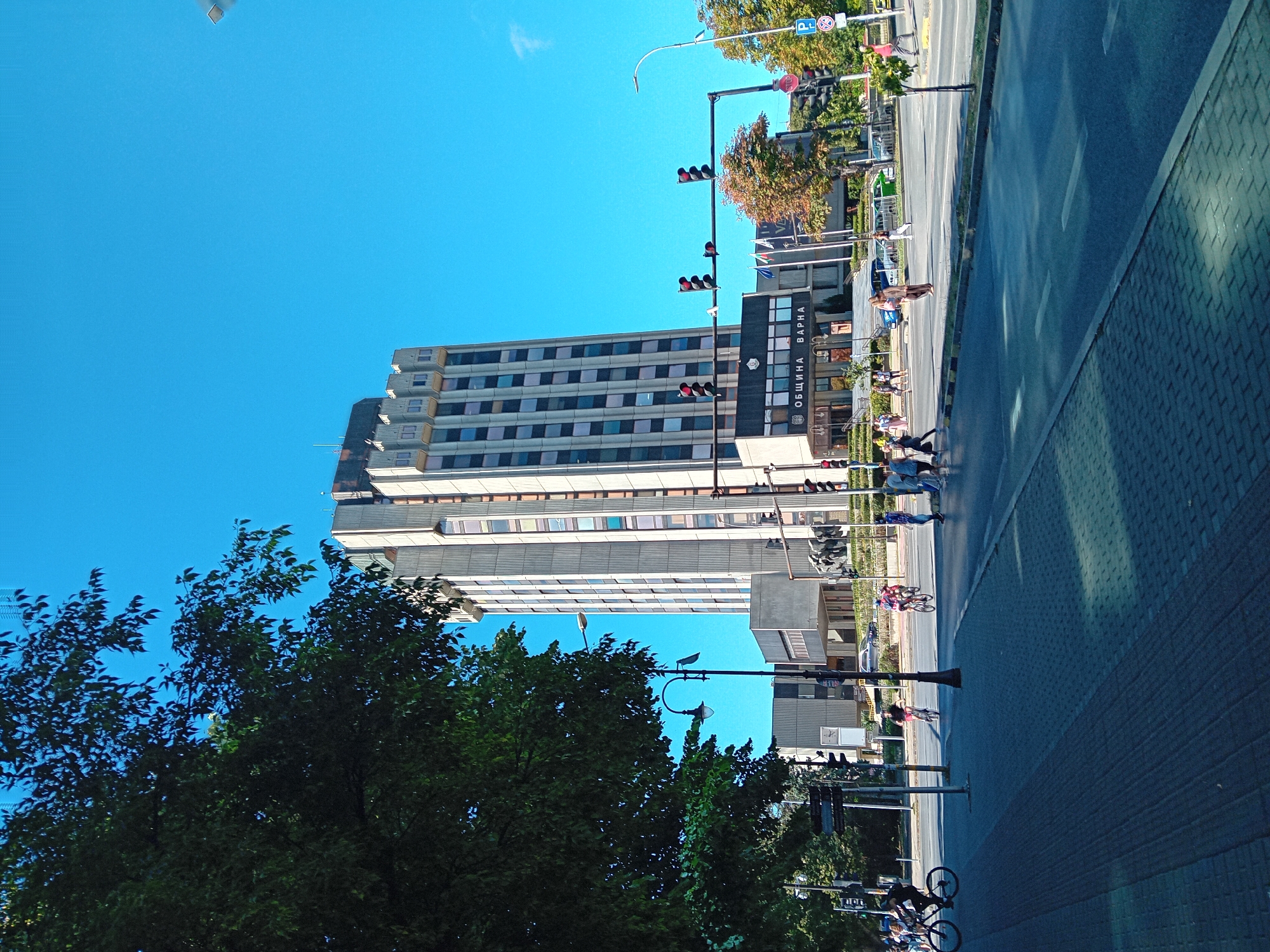 The image shows a street scene in Varna, Bulgaria, with the Municipality of Varna building prominently featured. A tall, concrete building with numerous windows dominates the background. In the foreground, a tree on the left partially obscures the view. A paved sidewalk runs alongside a road where several people are walking and cycling, and cars are visible. Traffic lights are present, indicating an intersection. The sky is clear and blue, suggesting a sunny day.