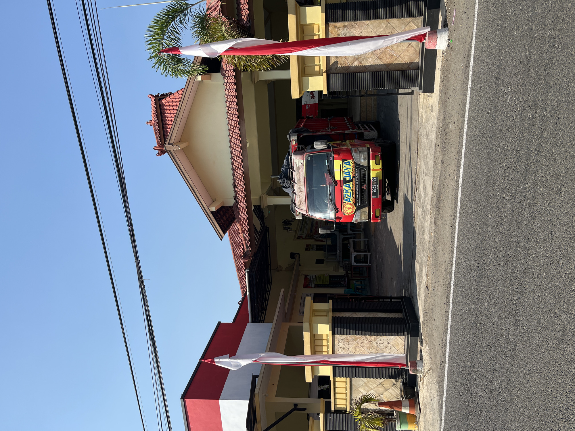 The image depicts a scene in front of a building with a red and white facade, located on a street in Margorejo, Indonesia. A red truck with the branding "AZKA JAYA" is parked in front of the building. Two Indonesian flags, red and white, are prominently displayed on flagpoles. The building has a tiled roof and a covered entryway. A palm tree is visible to the right of the flags. The street in the foreground is paved with asphalt and has a white line marking. The sky is clear and blue. Several potted plants and traffic cones are present near the entrance of the building. The overall impression is that of a commercial or administrative building on a sunny day.