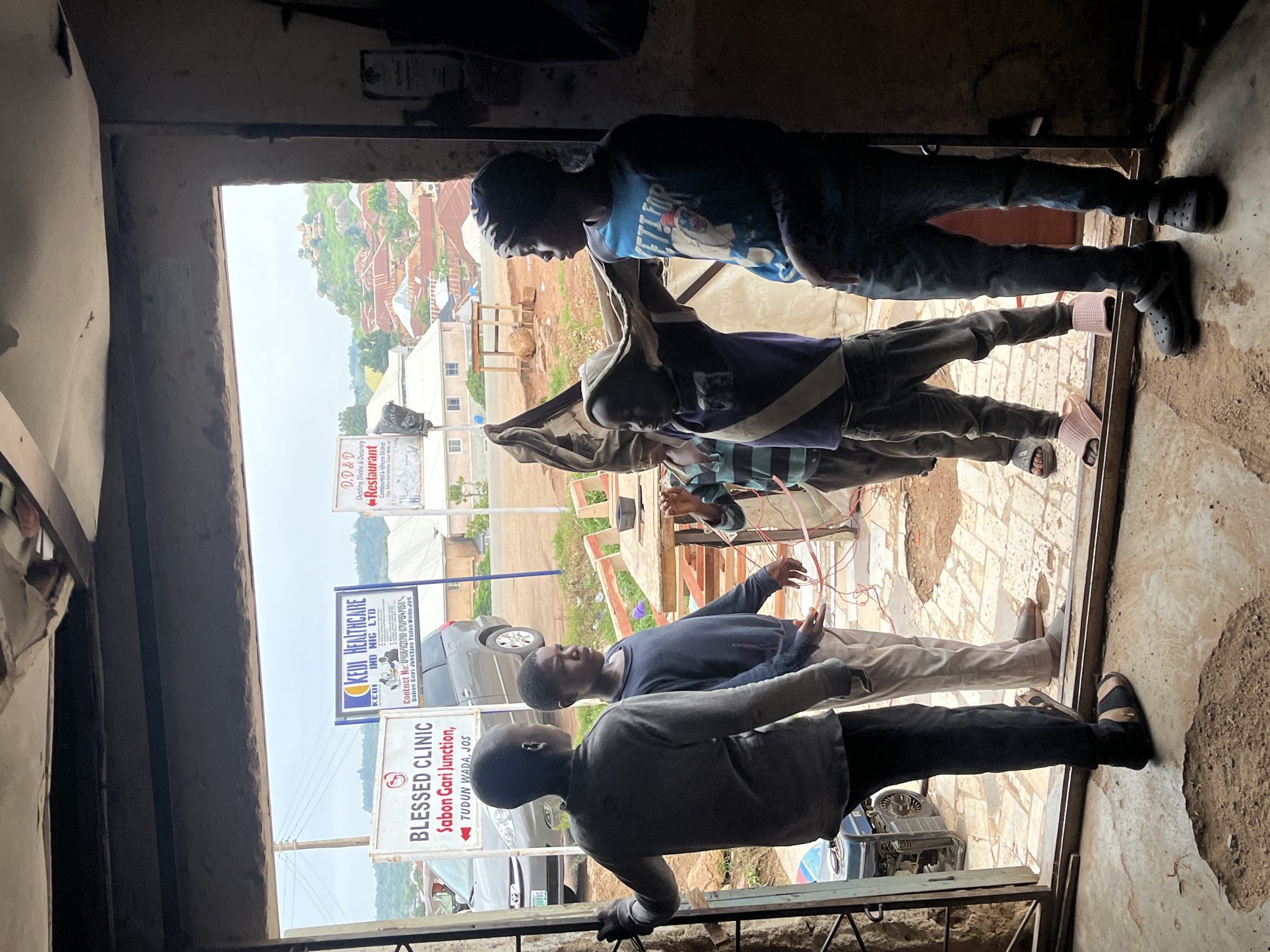 The image depicts a street-level view from inside a doorway looking outwards towards a scene in Jos, Nigeria. The foreground shows the lower legs and feet of four young males. The environment outside appears to be a commercial area with signs indicating local businesses. One sign reads "BLESSED CLINIC Sabon Gari Junction, Tudun Wada, Jos." Another sign is for "KEDI HEALTHCARE IND. NIG. LTD." and provides a contact number and location. A third sign appears to be for a restaurant. The background shows a hilly landscape with numerous buildings with red-tiled roofs, characteristic of Jos. A grey vehicle is parked near the "Blessed Clinic" sign. The ground inside the doorway is rough concrete with visible holes and debris. The overall lighting suggests it is daytime.