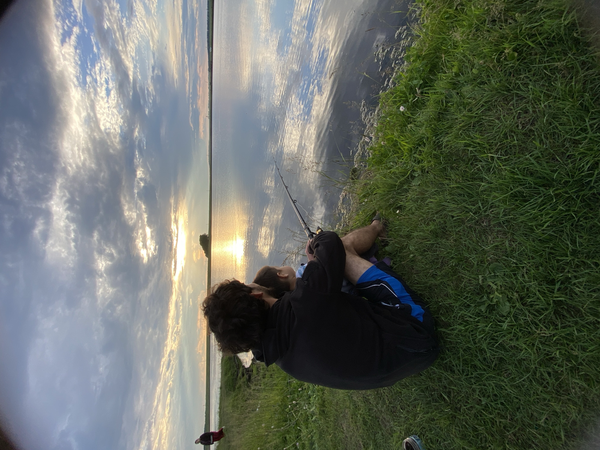 The image captures a serene sunset scene by a lake in Сыйрышбашево, Russia. In the foreground, a person with dark, curly hair wearing a black hoodie and blue shorts sits on the grass, facing away from the camera. They are holding a fishing rod, with their arm around a young child seated in front of them. The child is wearing a light-colored shirt. The lake's surface reflects the dramatic sky, which is filled with a mix of dark and bright clouds, with the sun's rays peeking through. In the distance, a thin strip of land with some trees and structures is visible. To the left, another person is walking along the grassy bank. The environment is rural, characterized by the lake, abundant grass, and a peaceful sky.