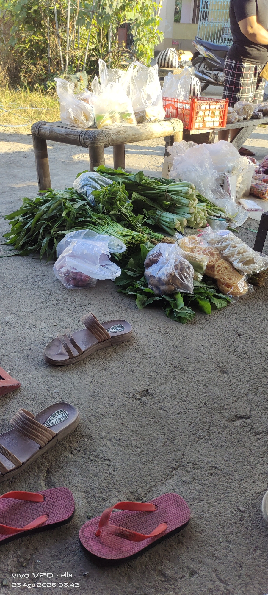 The image shows a ground-level view of a market stall or roadside stand in Simo, Indonesia. In the foreground, a pair of brown sandals are placed on a concrete surface, with another pair of pink flip-flops to the lower left. Behind the sandals, a large bunch of leafy green vegetables, possibly water spinach or kangkong, is spread out. Several plastic bags containing produce and other items are visible. One bag contains what appear to be red onions. Another bag contains fried snacks, such as crackers or crisps, and a bag further to the right contains what looks like ginger or other root vegetables. In the background, a wooden bench or table is set up, also holding bags of goods, some in plastic and some in a red plastic crate. A motorcycle is parked behind the stall, and a person wearing a dark shirt and checkered pants is partially visible on the right side of the image, suggesting they are operating the stall. The scene appears to be outdoors, with sunlight casting shadows and illuminating the vegetation in the background. The overall impression is a typical scene at a local Indonesian market.