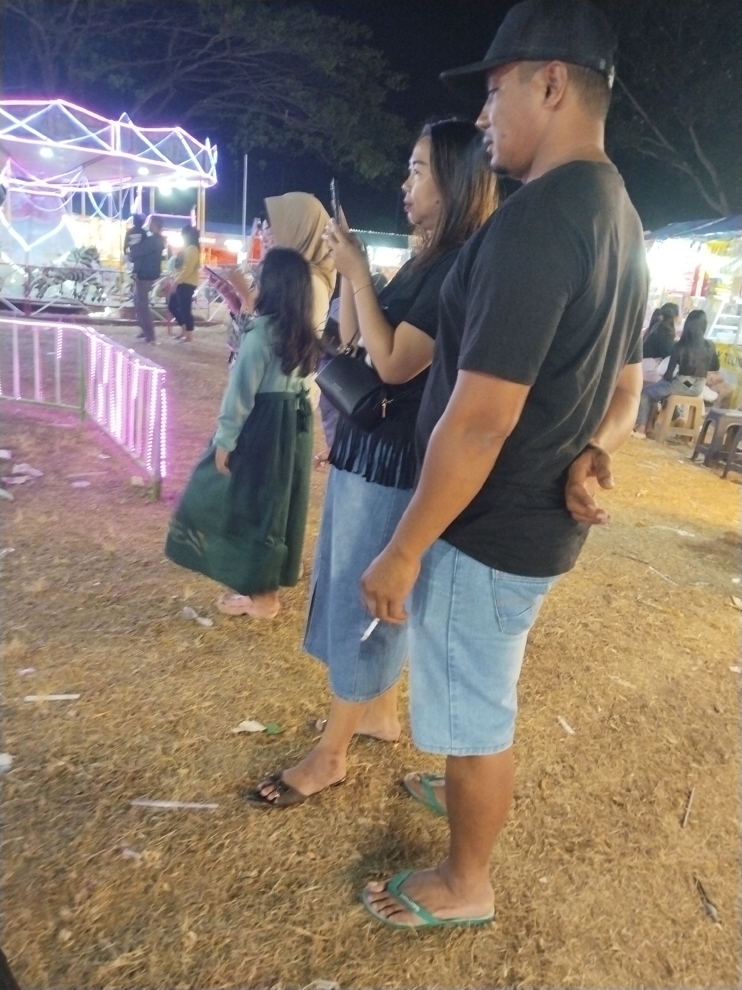 At night in an outdoor event space in Tasikmadu, Indonesia, two adults and two children are visible. A man wearing a black baseball cap, black t-shirt, and light blue denim shorts stands on the right side of the frame, facing left. He has his left hand behind his back and is holding a cigarette in his right hand. Next to him, a woman wearing a black t-shirt and a light blue skirt stands facing left. She is holding a phone in her right hand. To the left of the woman, a child wearing a dark green dress and pink sandals stands facing left. Behind them, another child wearing a light brown hijab and a green dress stands facing left. In the background, a brightly lit amusement ride with neon lights is visible, along with other people and a food stall. The ground is covered in dry grass and debris.