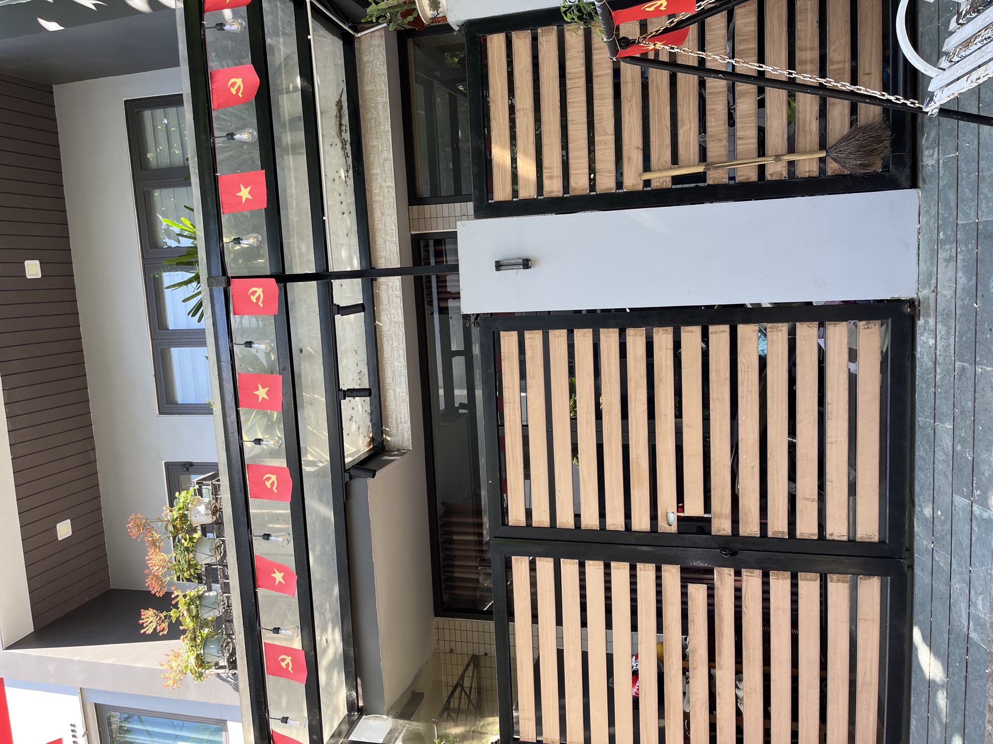 The image depicts a residential scene in Đà Nẵng, Vietnam. A house with a gray exterior and dark-framed windows is visible. A metal structure adorned with Vietnamese flags and light bulbs overhangs the entrance area. Potted plants are also present. A gate with a black frame and wooden slats sits to the right. A broom rests against the upper part of the gate. The ground is paved with gray stone tiles.
