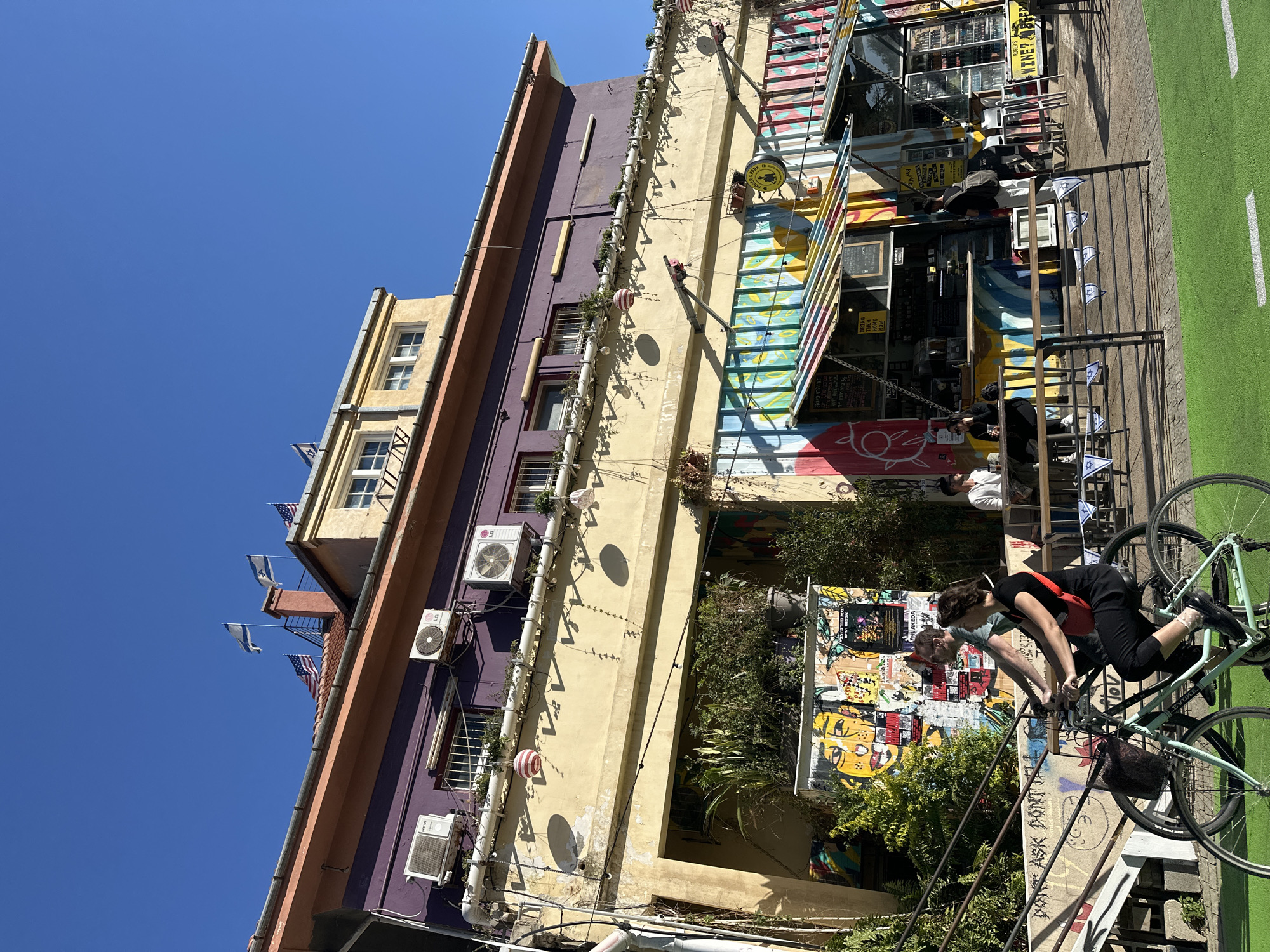 This vibrant, sunlit image captures a bustling street scene in Tel-Aviv, Israel, under a clear blue sky. Multi-story buildings with brightly colored facades in shades of purple, yellow, and orange line the street. Air conditioning units and decorative planters adorn the building walls.
In the foreground, two individuals are prominent: a woman in black pants and a red top is leaning over a light green bicycle, next to another person in a green shirt. Several other people are visible further down the street, seated at outdoor tables and chairs belonging to a cafe or bar setup, complete with colorful striped awnings.
The street features a distinct green-painted strip, likely a bike lane, running alongside paved pedestrian areas. Walls are decorated with a mix of street art, posters, and graffiti. Small Israeli flags (blue Star of David on white) and American flags are seen hanging from balconies, indicating the location.
Visible text includes phrases such as "BRING THEM HOME NOW" and "AKEDA" on posters, "ASK DON'T" on a wall, and business names like "FUMMY," "ROGER'S," and "WINE?" on shop signs. The bright sun casts strong shadows, suggesting midday conditions and clear weather, contributing to a lively and casual atmosphere.