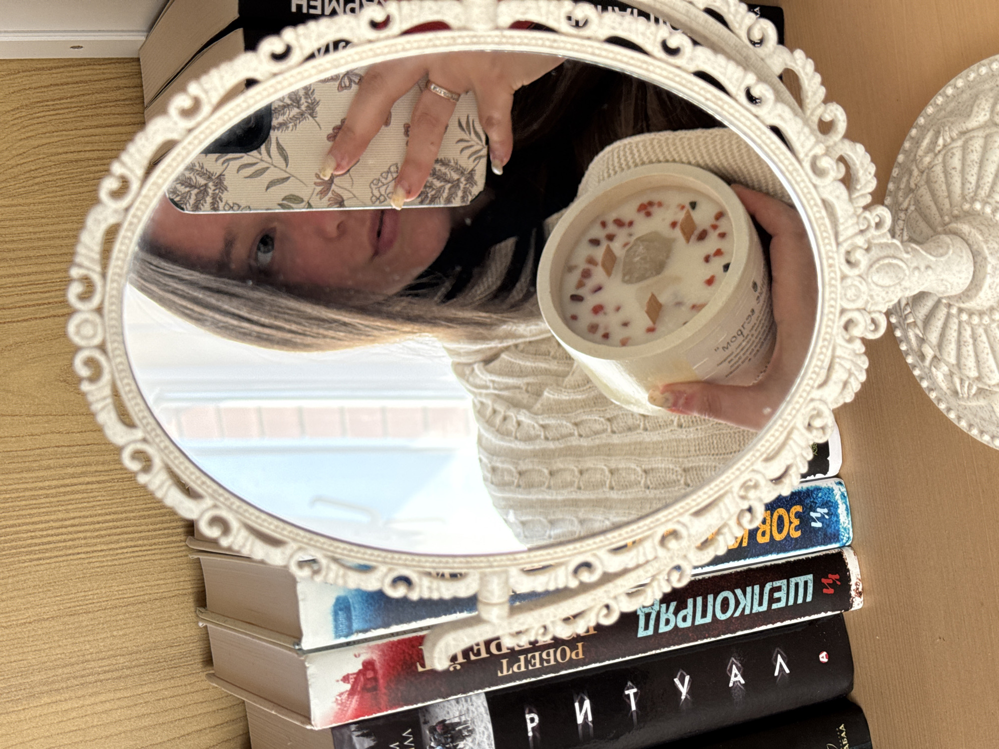An indoor mirror selfie captures a partial reflection of a person, likely female, holding a decorative candle and a smartphone. The oval-shaped mirror, featuring an ornate cream-colored frame, rests on a stack of books. The person, wearing a light-colored, possibly knitted, sweater, has light nail polish and a ring on one finger. Their smartphone, equipped with a botanical-patterned case, is held in one hand, while the other hand cradles a cream-colored candle adorned with red/pink fragments, small rectangular pieces, and a central clear stone or crystal.
The background shows a light-colored wooden surface, likely a table or shelf. Through the reflection in the mirror, a window and exterior building are visible, suggesting daytime. The books under the mirror are identified by their Cyrillic titles and author names, visible in reverse: "Р М Е Н", "ЗОВ", "ШЕЛКОПРЯД" (Silkworm), "РОБЕРТ" (Robert), and "Р И Т У А Л" (Ritual). A partial label on the candle shows "mogar..." or similar text, also reversed. This scene is set in Krasnodar, Russia.