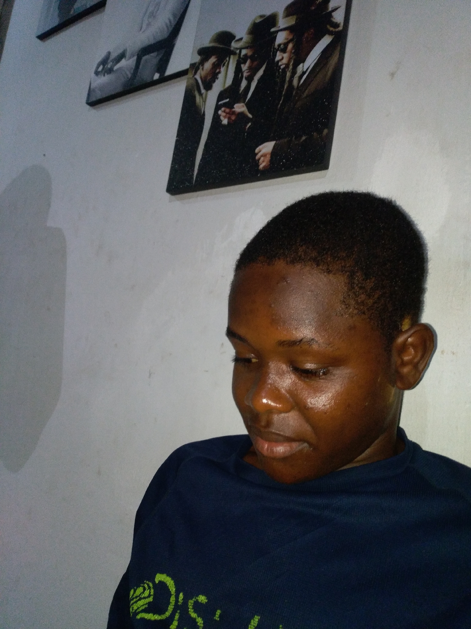 The image displays a head-and-shoulders portrait of a young individual with a dark complexion, positioned indoors against a plain, light-colored wall. The person's head is tilted downwards, with eyes appearing closed or cast towards the bottom right, suggesting a pensive or contemplative state. They have short, dark, closely-cropped hair. A sheen is visible on the forehead and nose, indicating either natural skin oils or direct lighting. The individual is wearing a dark blue crew-neck shirt, with a partial lime green graphic design visible on the left shoulder/sleeve.
On the wall behind the individual, three framed pictures or canvases are mounted. The most prominent one, positioned to the upper right, depicts three men dressed in suits and fedora hats, rendered in a classic or vintage style. Two other, less distinct framed artworks are visible higher and further to the left on the wall. The wall itself appears to be a light off-white or light gray, showing some minor texture and faint marks. A soft shadow of the individual's head and upper body is cast on the wall to the upper left. The specific geographic location within the city of Unknown, Unknown, cannot be determined from the visual information provided.