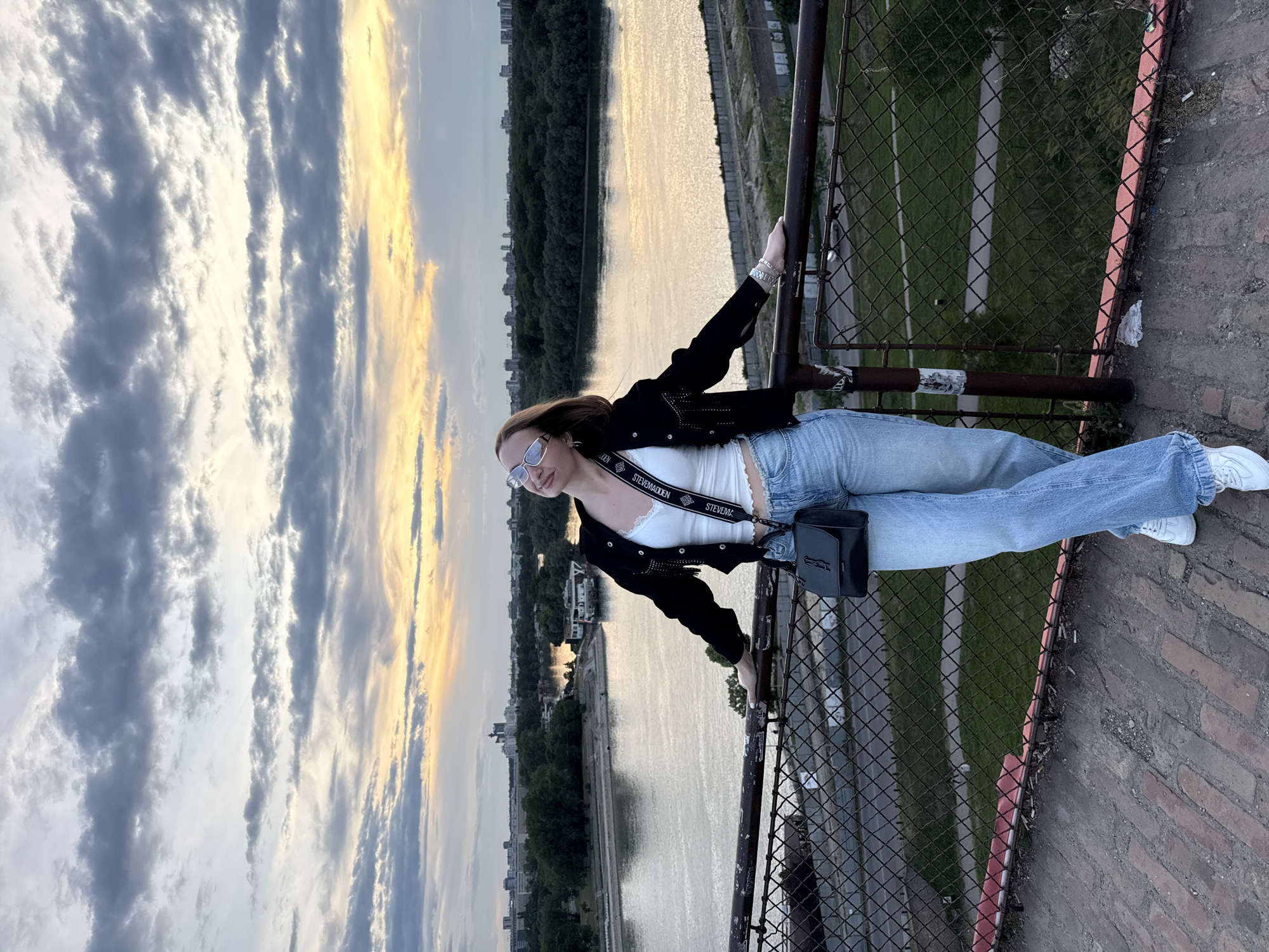 A woman in a white tank top, light-wash blue jeans, a black jacket, and white sneakers stands on a bridge or high platform overlooking a river. She is leaning against a rusty metal railing with a chain-link fence below. She is wearing sunglasses and has a black purse slung over her shoulder. The background shows a river with trees and buildings on the opposite bank. The sky is filled with clouds tinged with yellow, suggesting it is near sunset. The location is Belgrade, Serbia, identifiable by the landmarks and architectural styles.