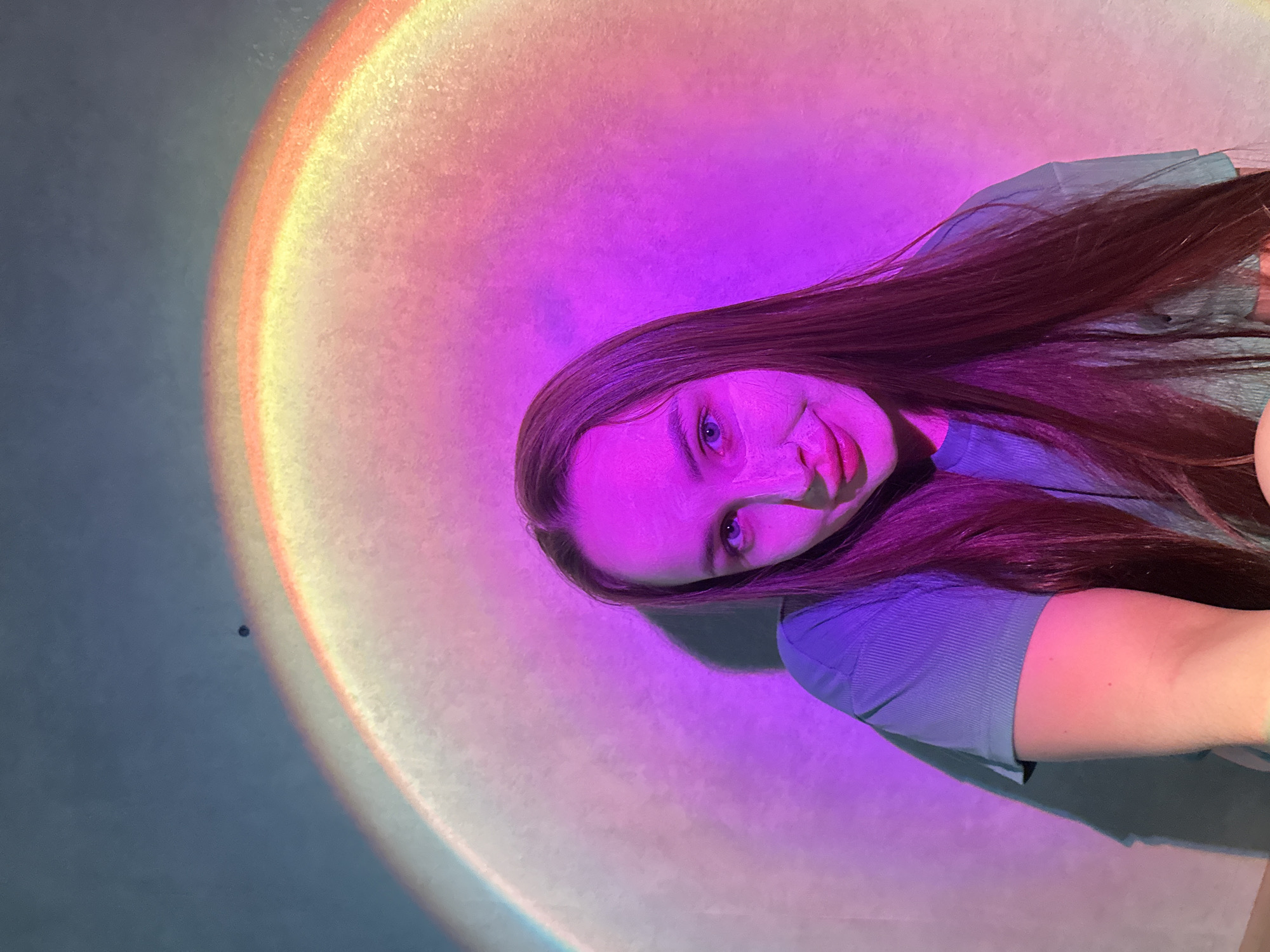 A young woman with long, dark brown hair is captured in a close-up, slightly rotated portrait. She looks directly at the camera with a subtle smile. Her face and part of her hair are bathed in vibrant magenta and purple light, casting a glow that accentuates her features and blue eyes. She is wearing a ribbed, blue-grey short-sleeved top.
The background is dominated by a large, circular, gradient light projection, resembling a sunset lamp effect. This projection transitions from bright yellow and orange at its outer edge, through shades of warm pink, to a deeper magenta and purple closer to the woman. A portion of the background to the far left of the light is a muted grey-blue. A small, dark, circular speck is visible in the grey-blue area of the background. The overall setting appears to be indoors, likely in a room, with the artificial light source creating a distinct ambiance. There is no visible text. The location is Chaikovsky, Russia.
