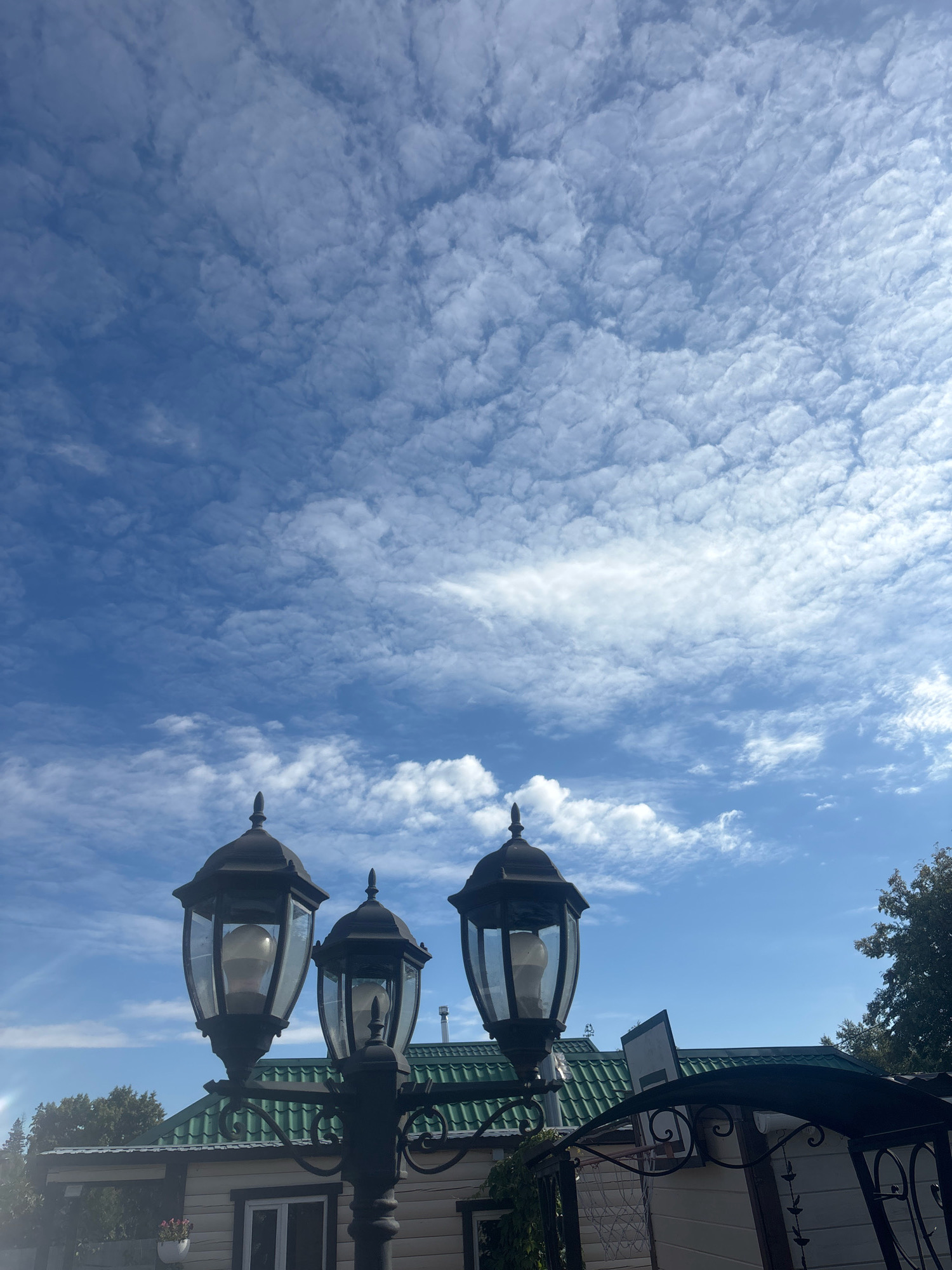 In Лотос Снт (марьино), Russia, this image captures an outdoor scene dominated by a bright blue sky filled with scattered, wispy white clouds. In the foreground, three ornate, black lampposts with glass enclosures are visible, each housing a light bulb. Behind the lampposts, a building with a green, tiled roof and light-colored siding is partially visible. The building has a traditional aesthetic, with visible window frames. To the right of the building, the branches of a leafy green tree extend into the frame. The overall impression is a serene, daytime view of a residential or recreational area.