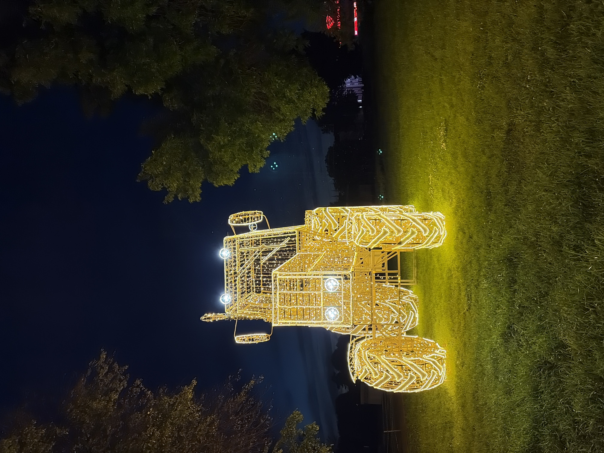 The image displays a prominent, brightly illuminated sculpture of a tractor at night. The tractor, constructed from a framework covered in numerous yellow and golden lights, emanates a strong glow. Its design includes distinct large wheels, a cabin, and what appears to be a front-mounted attachment, possibly a plow or loader. Several brighter, circular lights are integrated into the sculpture, acting as headlights and other decorative elements.
The sculpture stands on a grassy area, which is bathed in a yellowish-green light reflecting from the tractor. In the background, dark trees with dense foliage are silhouetted against a deep blue-black night sky. Faint blurs of light in the distant background suggest other buildings or ambient city lights.
The scene takes place at night under a clear sky. There are no people visible in the image. This appears to be an outdoor light art installation, likely part of a festive or public display in Minsk, Belarus. The illuminated tractor evokes a sense of wonder and visual spectacle. No visible text is present in the image.