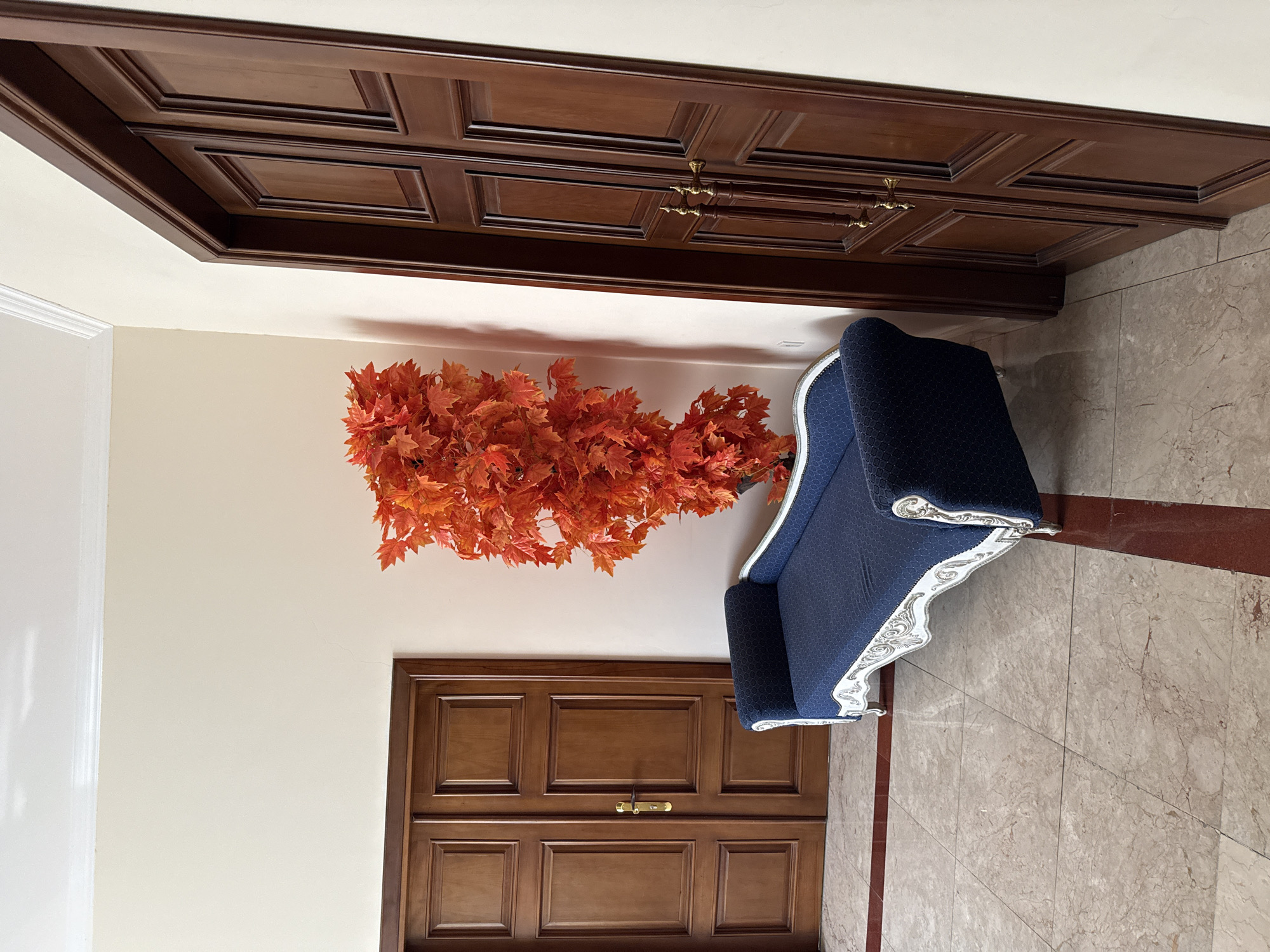 The image depicts an interior scene with a blue chaise lounge and an artificial tree with bright orange leaves. The chaise lounge is ornate, with white carved details and a patterned blue fabric. It is positioned on a marble floor with dark red inlaid stripes. Behind the chaise lounge, two tall, dark wooden double doors are visible, one on the left and one on the right. The door on the left has a gold handle, while the door on the right has two gold handles. The walls are painted a light, neutral color. The scene appears to be set in a hallway or entrance area of a building. Based on the provided location, this scene is likely situated within a residential or commercial property in Sabail Raion, Azerbaijan. There are no people present in the image.