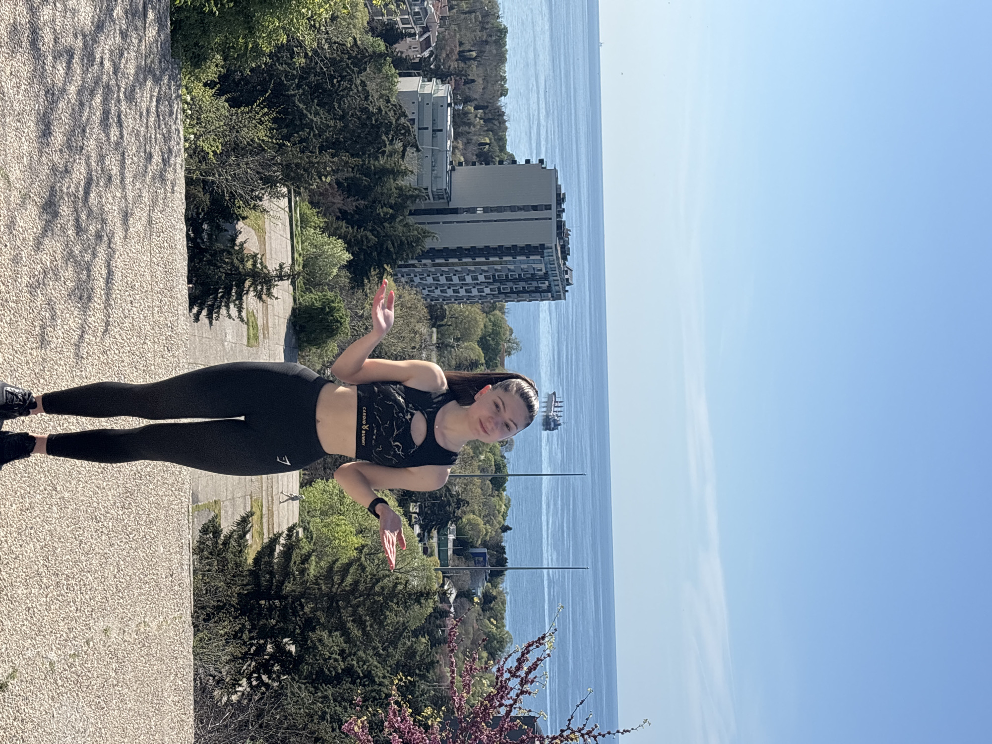 The image captures a young woman standing outdoors on what appears to be an elevated vantage point, overlooking a coastal scene. She is positioned on a light-colored, pebbled surface, facing the camera with a subtle smile. Her hands are slightly raised, suggesting a casual pose or wave.
The woman is dressed in athletic attire: a black sports bra with a pattern and a cut-out detail, displaying the text "CARDIO BUNNY" on the band. She also wears black leggings, on which a white logo resembling the Gymshark shark is visible on her left thigh, and black athletic shoes.
Behind her, the landscape unfolds with lush green trees and foliage. In the mid-ground, a tall, multi-story building, possibly a hotel or apartment complex, stands prominently near the water's edge. Beyond the building and trees, an expansive body of water, likely the sea or ocean, stretches towards the horizon under a clear, bright blue sky with a few faint wispy clouds. A ship or boat can be seen in the distance on the water. In the lower right corner of the scene, there are flowering bushes with purple blossoms, and two tall, slender poles or masts rise from the greenery.
The time of day appears to be bright daylight, with strong sunlight indicating clear weather. The overall impression is one of a relaxed pose in a scenic, sunny, coastal environment.