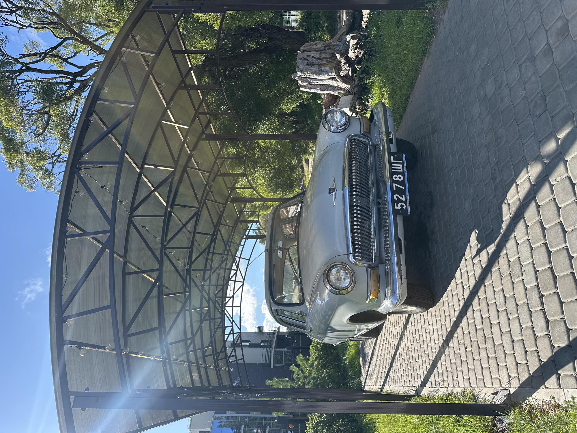 A vintage, light grey GAZ-21 Volga sedan is parked under a covered carport with a transparent roof and a dark metal frame. The car faces the viewer, positioned centrally on a paved driveway made of grey interlocking stones. The car's license plate reads "5278 ШГ". To the right of the car, a large, weathered tree stump is visible, surrounded by grass and more trees. In the background to the left, a modern multi-story building with dark and light facades is partially visible, along with greenery and a blue sky with scattered clouds. The scene suggests an outdoor setting, possibly in a residential or park area, on a sunny day.