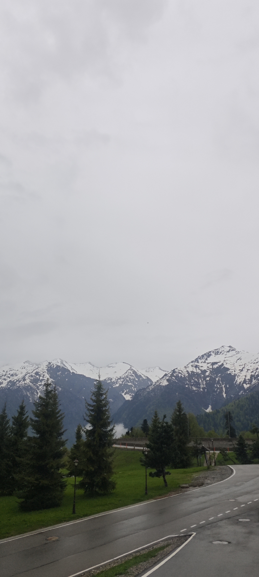 The scene is a landscape in Unknown, Russia, featuring a foreground with a wet asphalt road with white lane markings, curving slightly to the right. To the left of the road, there is a grassy area dotted with several coniferous trees and short, dark lampposts.
In the background, a mountain range is visible, with snow-capped peaks under a gray, overcast sky. The mountains are partially obscured by fog or low-hanging clouds. The overall environment suggests a cool, possibly wet or recently rainy day in a mountainous region. There are no people visible.