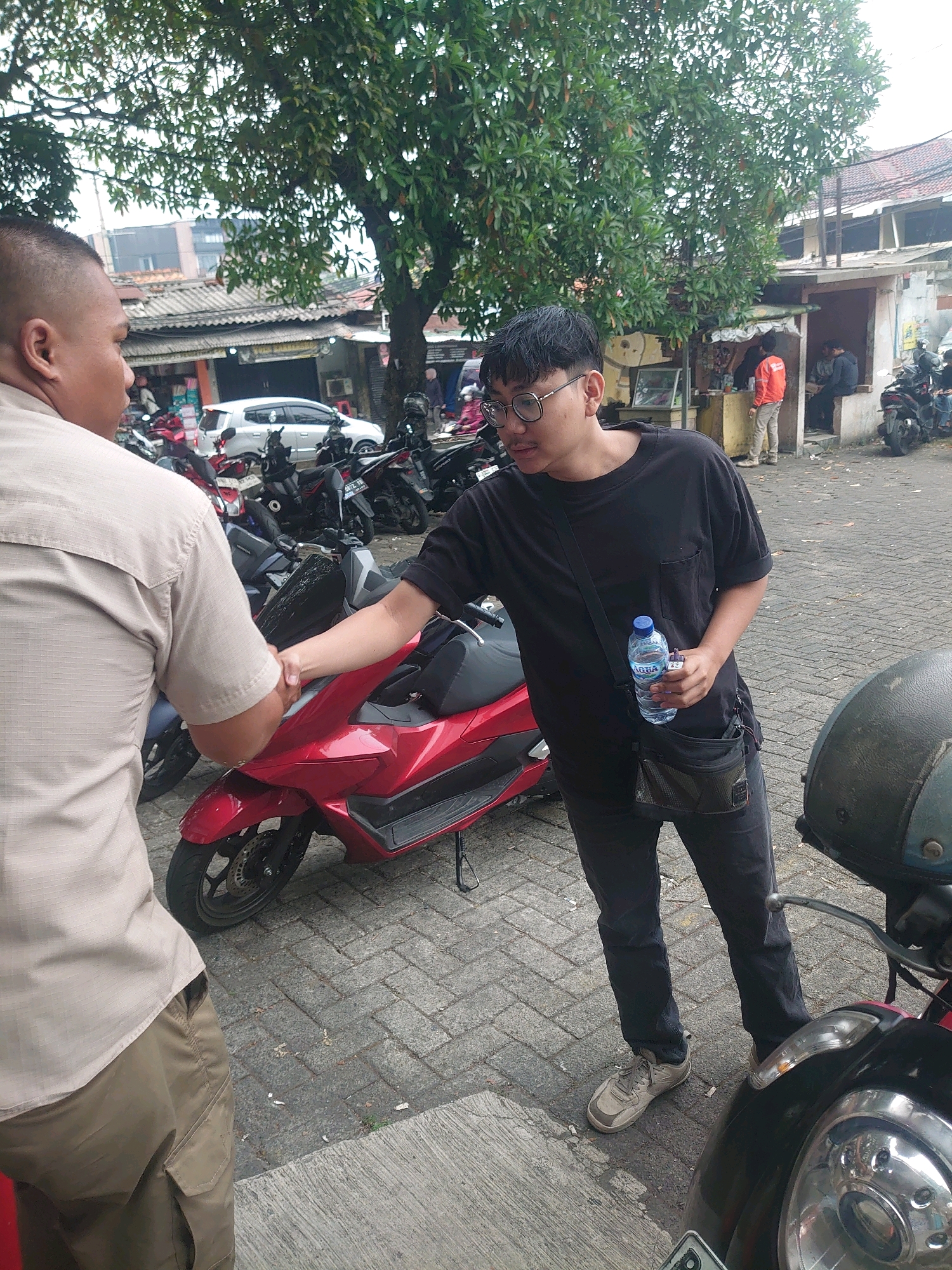 In South Jakarta, Indonesia, two men are seen shaking hands in what appears to be an outdoor marketplace or parking area. The man on the left, wearing a tan short-sleeved button-up shirt and khaki cargo pants, has his back to the camera. The man on the right, wearing a black t-shirt, black pants, and glasses, is holding a bottle of Aqua water in his right hand and shaking the other man's hand with his left. He also has a black crossbody bag slung over his shoulder. In the background, several parked motorcycles, including a red scooter in the foreground, are visible, along with stalls and buildings typical of an urban commercial area. A large tree with green foliage provides shade. The ground is paved with interlocking bricks.