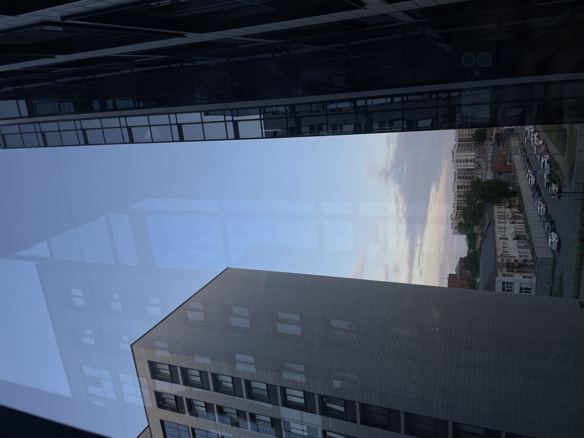 The image depicts a view from a window in Sinyavino, Russia, showcasing a juxtaposition of modern architecture and older buildings. In the foreground, two large buildings dominate the frame. The building on the left is a multi-story structure with a light-gray, textured facade and numerous dark-framed windows. Its corner is sharply angled, emphasizing its contemporary design. To the right, a darker, sleek building rises, characterized by a facade of dark glass panels arranged in a grid. Reflections of the sky and other structures are visible on its surface.
Beyond these prominent buildings, the scene transitions to a view of the city. A strip of older, lower-rise buildings with light-colored walls and numerous windows stretches across the middle ground. A street runs alongside these buildings, lined with parked cars of various colors, including white, black, and silver. Trees and greenery are interspersed between the buildings, suggesting a moderately developed urban environment. The sky above is a gradient of blue and pale orange, with wispy clouds, indicating either dawn or dusk. The overall impression is one of urban development with a mix of architectural styles.