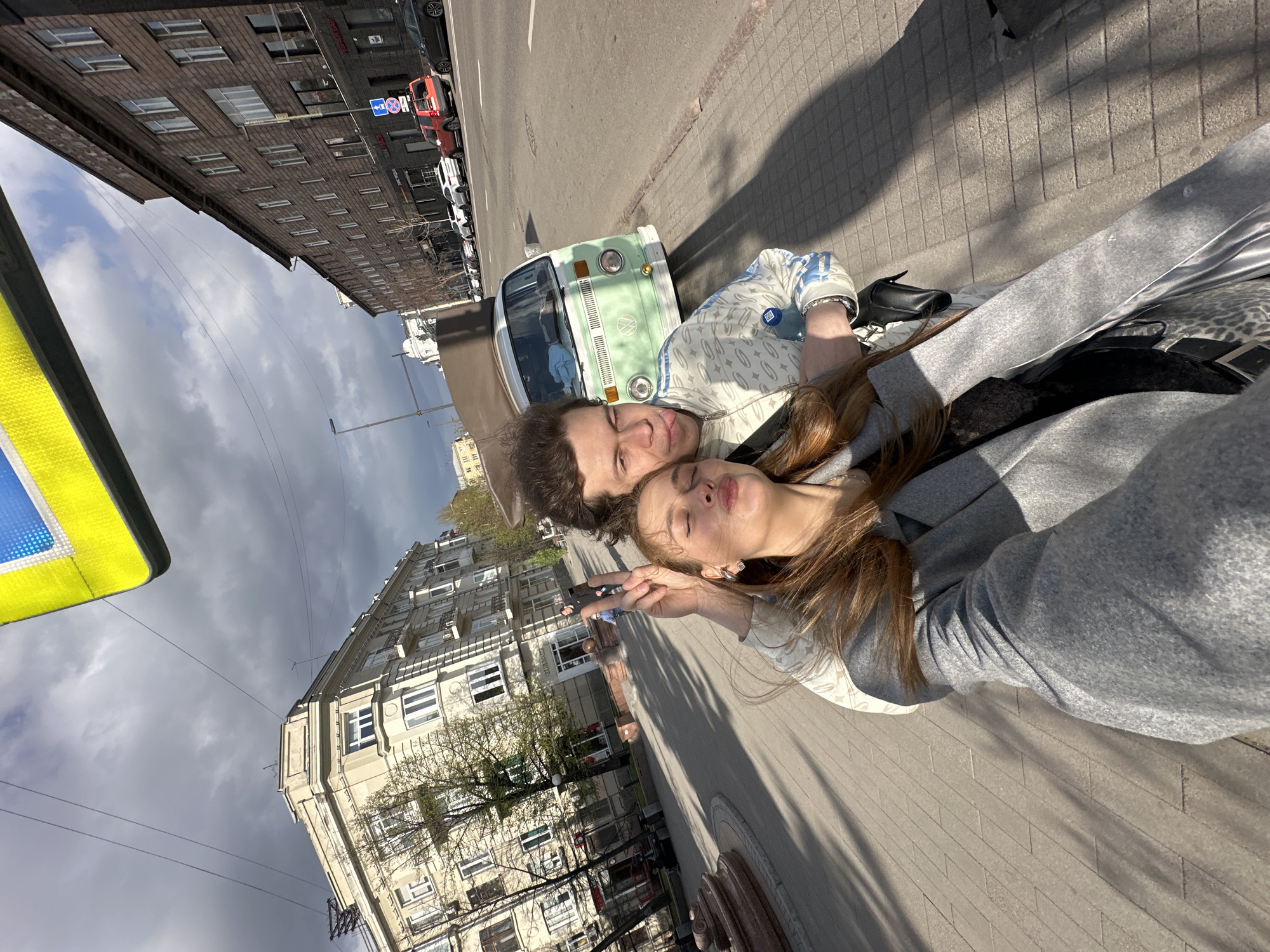 Two people, one male and one female, are posing for a selfie. The woman has long brown hair and is making a peace sign with her fingers. The man has short brown hair. Behind them, there is a light green Volkswagen van. The photo appears to be taken in the city of Krasnoyarsk, Russia, evidenced by the buildings in the background. The street is paved with cobblestones. Other cars are parked on the street. The sky is cloudy. A yellow and blue sign is visible in the lower left corner.
