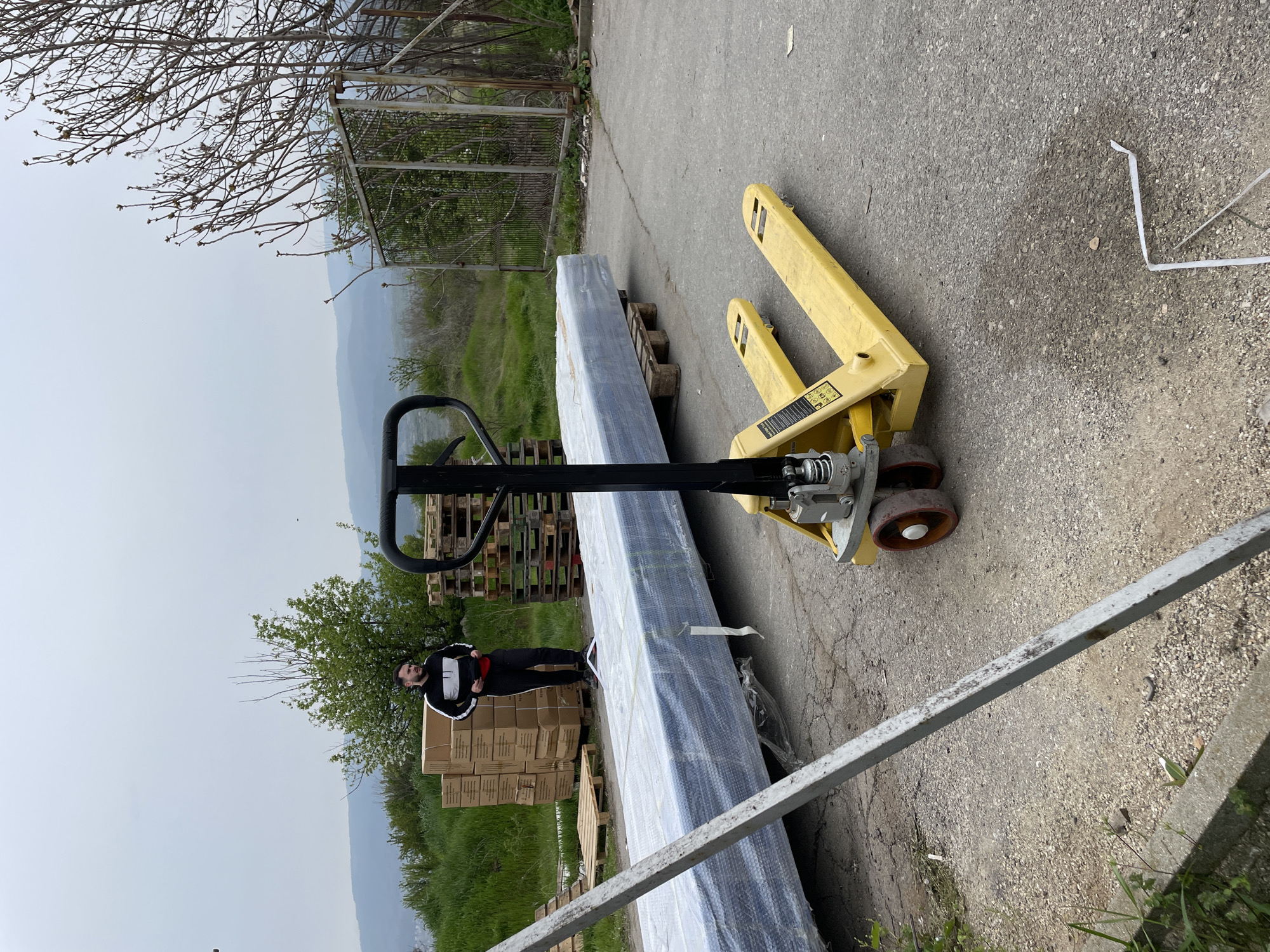 The image shows a scene outside in what appears to be an industrial or commercial area. A yellow pallet jack sits on a gray asphalt surface. The jack supports a large, wrapped item, likely merchandise or materials, resting on a wooden pallet. Nearby, there are stacks of cardboard boxes and additional pallets. A man wearing a black and white jacket is standing to the left of the wrapped object. In the background, there is green vegetation, including grass and trees, with a fence partially visible. The location is Asenovgrad, Bulgaria.