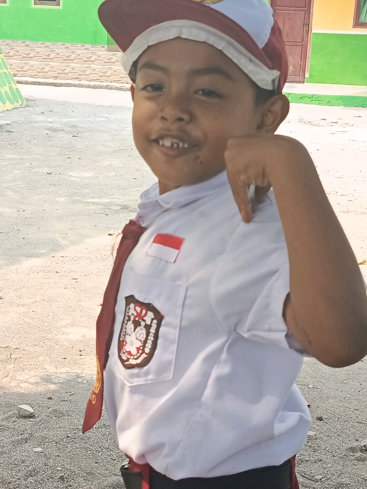 The image shows a young boy in a school uniform in Karya Dadi, Indonesia. He is wearing a white shirt with a red and white flag patch on the left chest and a brown and white emblem on the right chest. A maroon scarf is draped around his neck and down the front of his shirt. He also wears a maroon and white cap. The boy is looking at the camera and smiling, with his left hand raised to his face, index finger extended. He appears to be outdoors, with a sandy or gravel ground and a building with a green wall and brown door in the background.