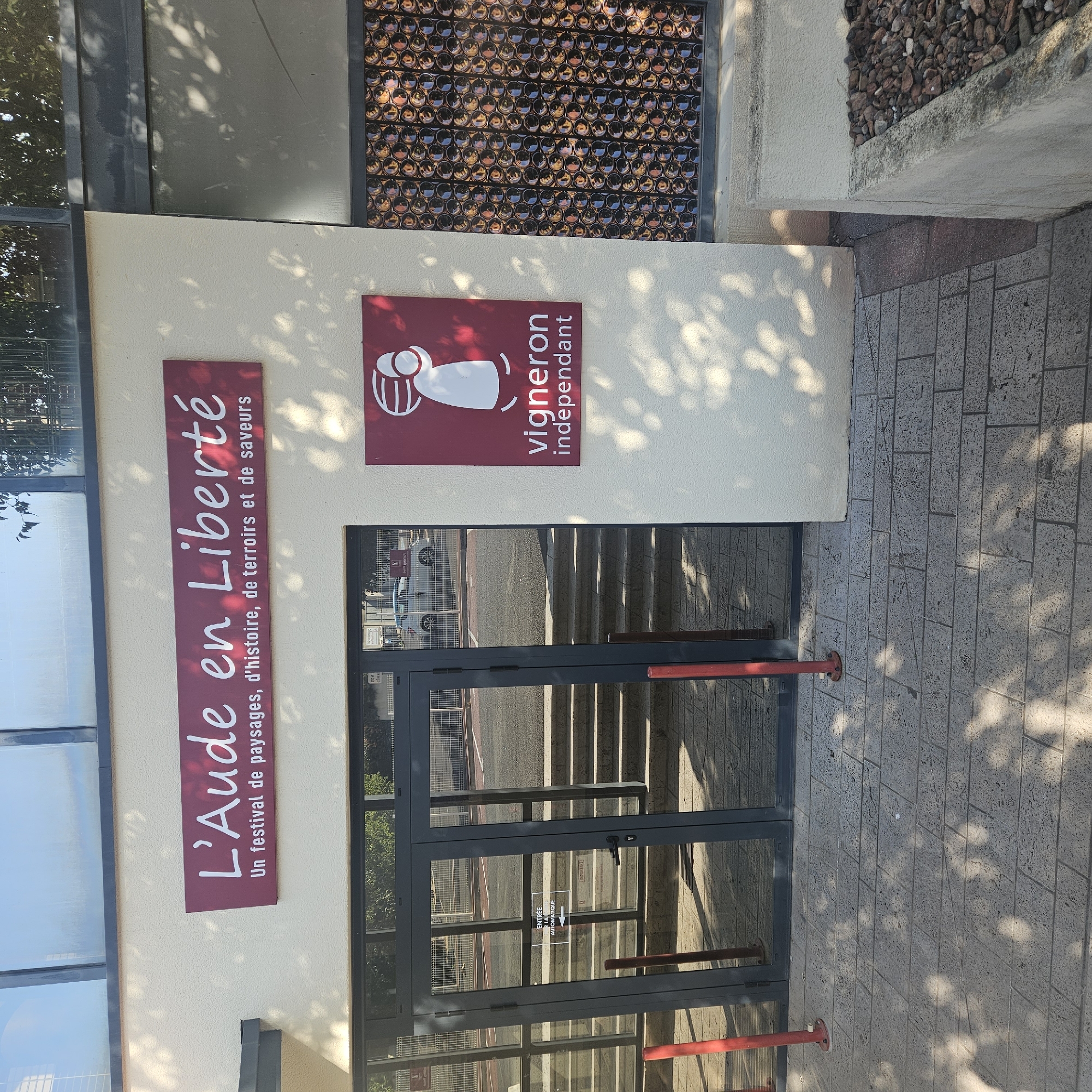 The image shows the exterior of a building with a sign that reads "L'Aude en Liberté" followed by the text "Un festival de paysages, d'histoire, de terroirs et de saveurs." To the right of this sign, there is a smaller square sign with a stylized white figure on a maroon background, above the text "vigneron indépendant." The building features glass doors with a sign on one of them indicating "ENTRÉE À LA AUTOMATIQUE" with an arrow. In the background, through the glass doors, a set of stairs and a paved area are visible, along with a white car and fencing. There are also decorative windows made of what appear to be glass bottles. The scene is illuminated by sunlight, casting shadows from trees. The building is located in Narbonne, France, as indicated by the sign. The overall impression is of an entrance to a cultural or commercial establishment.