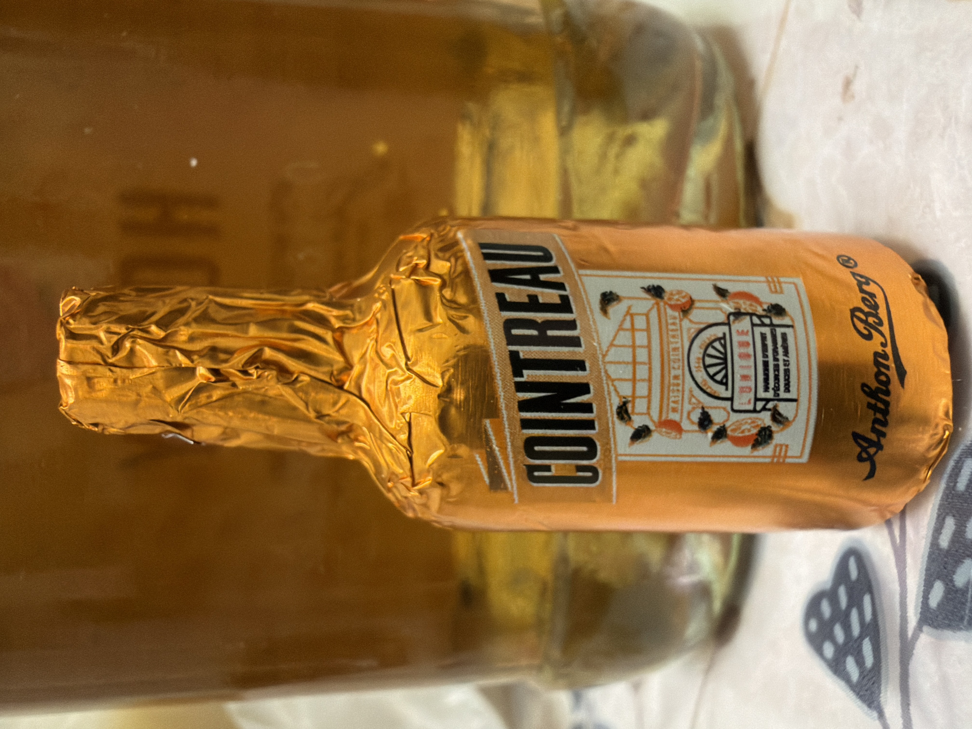 The image shows a close-up of a small, foil-wrapped bottle of Cointreau liqueur, likely a chocolate liqueur, branded by Anthon Berg. The bottle is predominantly orange and gold, with visible text that includes "COINTREAU", "UNIQUE", and "Anthon Berg". The bottle is placed on a patterned surface, possibly a tablecloth, with a blurred background of what appears to be a larger glass container filled with an amber-colored liquid, possibly more liqueur or a beverage. The lighting is warm, casting soft shadows. The scene is indoors, on a flat surface, with a cluttered background. The location in Kyiv, Ukraine, is not discernible from the image itself, as there are no specific landmarks or contextual clues to suggest this.