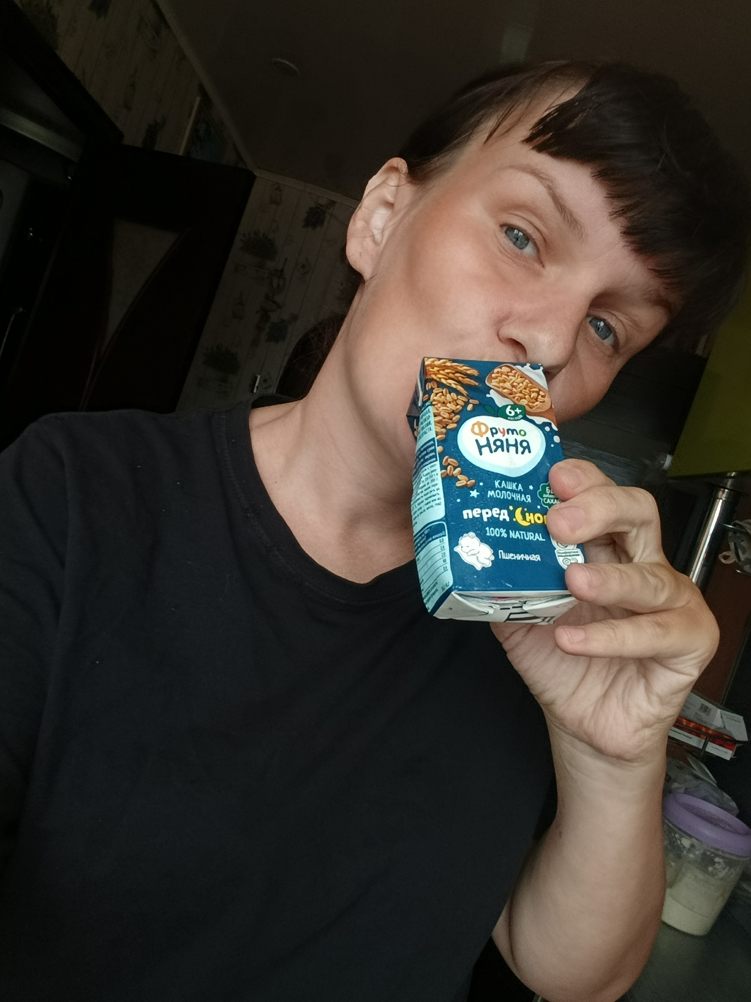 A woman is depicted in a selfie, holding a light blue cardboard drink carton to her mouth as if drinking from it. She has short, dark brown hair with bangs and striking blue eyes, looking directly at the camera with a neutral to slightly smiling expression. She is wearing a simple black t-shirt. The setting appears to be indoors, likely a home, with a dimly lit background featuring dark furniture or cabinetry and patterned wallpaper.
The carton is for "Фруто Няня" (Fruto Nyanya), a brand typically associated with baby food. Visible text on the packaging includes "6+" (indicating age suitability), "КАШКА МОЛОЧНАЯ" (Milk Porridge), "перед Сном" (before Sleep), "100% NATURAL", and "Пшеничная" (Wheat). "CAXAC" (Sugar) is also visible, possibly indicating its presence or absence.
The image suggests a casual moment, perhaps consuming a comforting beverage or snack before sleep, in a domestic environment in Фоки, Russia.