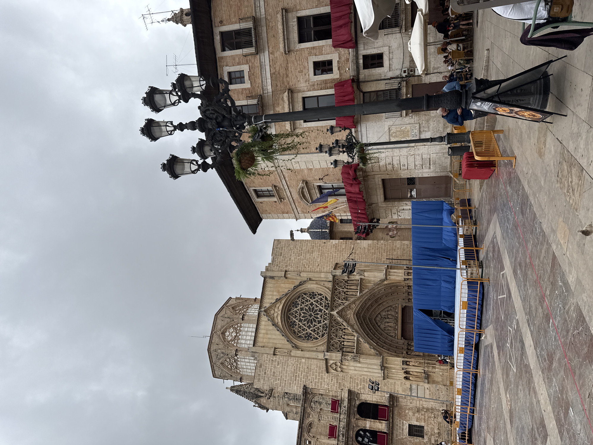This image depicts an urban square in València, Spain, under an overcast sky, showcasing historical architecture alongside modern temporary installations.
On the left side of the frame, a multi-story building with a light-colored brick or stone facade features several windows, some with dark frames and others accented by red fabric awnings. A large, ornate black metal street lamp with multiple glass lanterns and a hanging plant basket stands prominently in front of this building. Behind it, a distinctive blue and white tiled dome is visible, accompanied by flags, one of which resembles the regional flag of Valencia. An outdoor cafe area with white umbrellas, tables, and chairs is situated in the foreground on this side, occupied by several people. A man in a dark blue jacket stands near the street lamp, attending to a black A-frame sign that advertises "PIZZAS" and potentially "DELIVERY."
The right side of the image is dominated by a grand, historic Gothic-style building, likely a cathedral or basilica. Its intricate stone facade features detailed carvings, arched entrances, and a prominent central rose window with elaborate tracery, flanked by smaller circular windows. The lower part of this majestic structure is partially obscured by temporary blue fabric coverings and yellow metal crowd barriers, suggesting preparations for an event, performance, or ongoing construction/maintenance.
The ground of the square is paved with patterned stone tiles. The overcast sky indicates a cloudy day, making the exact time of day difficult to determine. The scene conveys a calm, functional atmosphere within a historical urban setting.