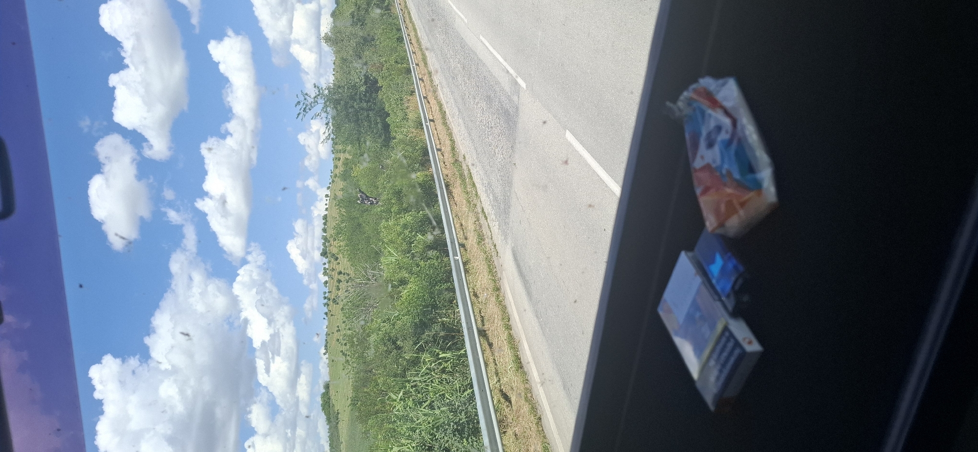 An exterior, eye-level, full shot captures a rural road view from inside a vehicle. The foreground features a dark dashboard with two packs of cigarettes and a small, blue item. The mid-ground displays a paved road with a white line, bordered by a silver guardrail and dry grass. Beyond the road, a lush green hillside is covered in trees and foliage, with a few scattered olive trees visible on the upper slopes. The sky above is a bright blue with scattered white cumulus clouds. In the distance, on the hillside, a person on a dirt bike can be seen. The location is specified as Maslarevo, Bulgaria.