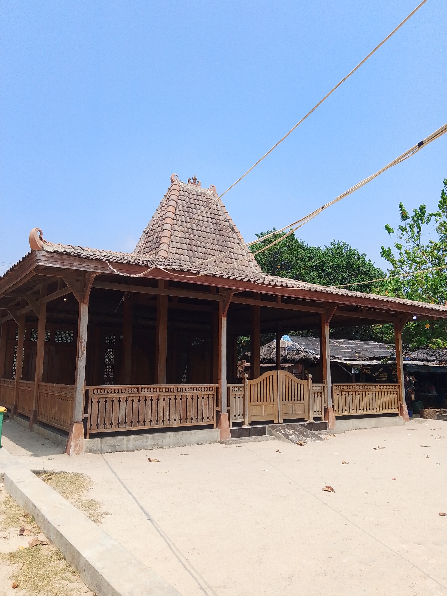 A traditional wooden pavilion with a tiled, multi-tiered roof stands under a clear blue sky in Pejagan, Indonesia. The structure, built on stilts, features intricate wooden latticework along its perimeter and a wooden gate. Several power lines are visible overhead, crossing the frame. The ground surrounding the pavilion is sandy, with scattered leaves. In the background, lush green trees and smaller structures with corrugated roofs are present. The scene is quiet, with no people or animals immediately visible.