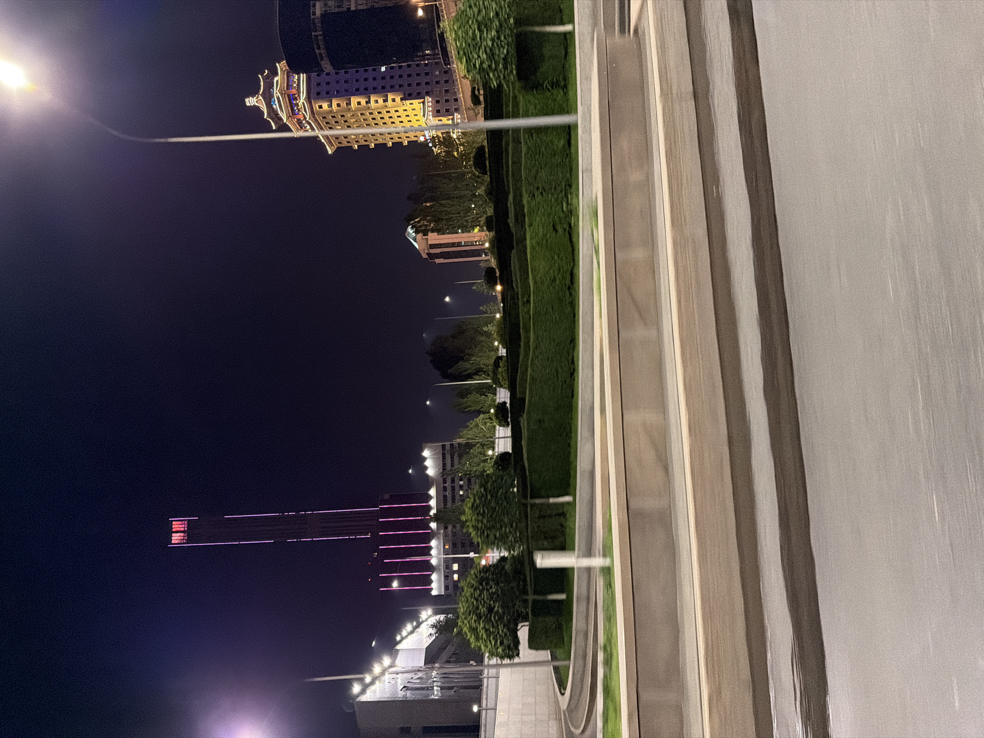 The image shows a nighttime view of a city street in Astana, Kazakhstan. The scene is dominated by modern architecture and artificial lighting, characteristic of an urban environment at night.
Prominent in the image are several tall buildings. One building, with a distinctive curved roofline and ornate facade, is illuminated with yellow and blue lights, giving it a grand appearance. Further down the street, other buildings are visible, some with rows of windows that are lit up, suggesting occupancy. A particularly striking building features vertical pink lights that highlight its structure against the dark sky.
A manicured green hedge runs along the side of the street, separating the buildings from what appears to be a pathway or walkway. Streetlights are visible, casting pools of light, and a bright streetlight illuminates the top of one of the buildings. The ground surface on the right side of the image is a textured, light-colored material that appears wet, possibly from recent rain or a cleaning process, and reflects the ambient light. The overall atmosphere is one of a well-lit, modern city at night. The dark sky suggests it is well past sunset, and the presence of lights and illuminated buildings indicates the city is active. There are no visible people or specific events taking place, only the static elements of the cityscape.