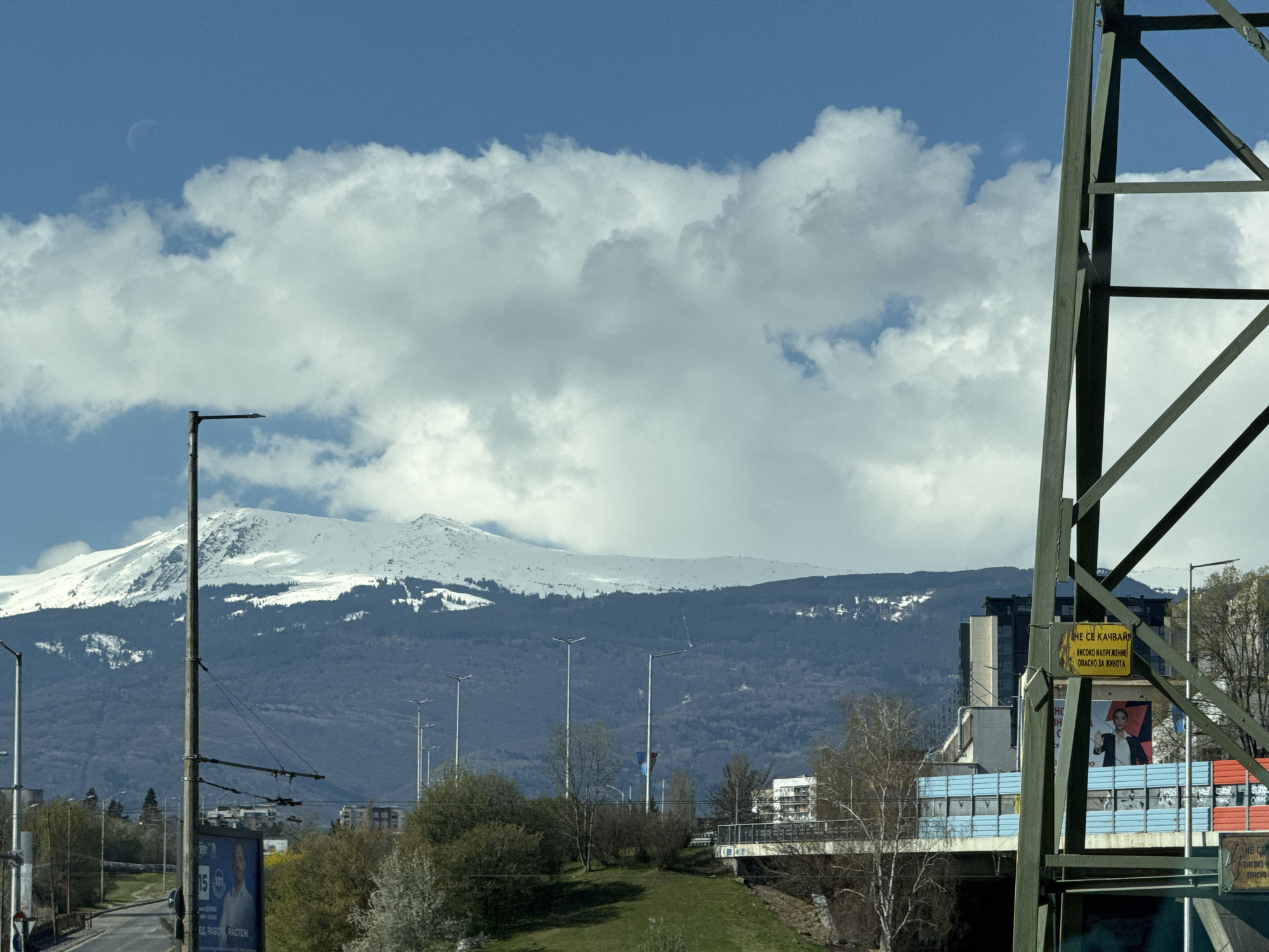 The image depicts a cityscape in Sofia, Bulgaria, featuring a view of Vitosha Mountain in the background, partially covered with snow. In the foreground, there's an electrical transmission tower to the right, obscuring some of the scene, and a street lamp on the left. A road is visible on the left-hand side of the frame. The foreground includes greenery and an overpass with blue railings and graffiti. A billboard is also visible. A sign attached to the electrical transmission tower has text in Cyrillic, warning about high voltage. The sky is blue with puffy white clouds, and a faint crescent moon is visible.